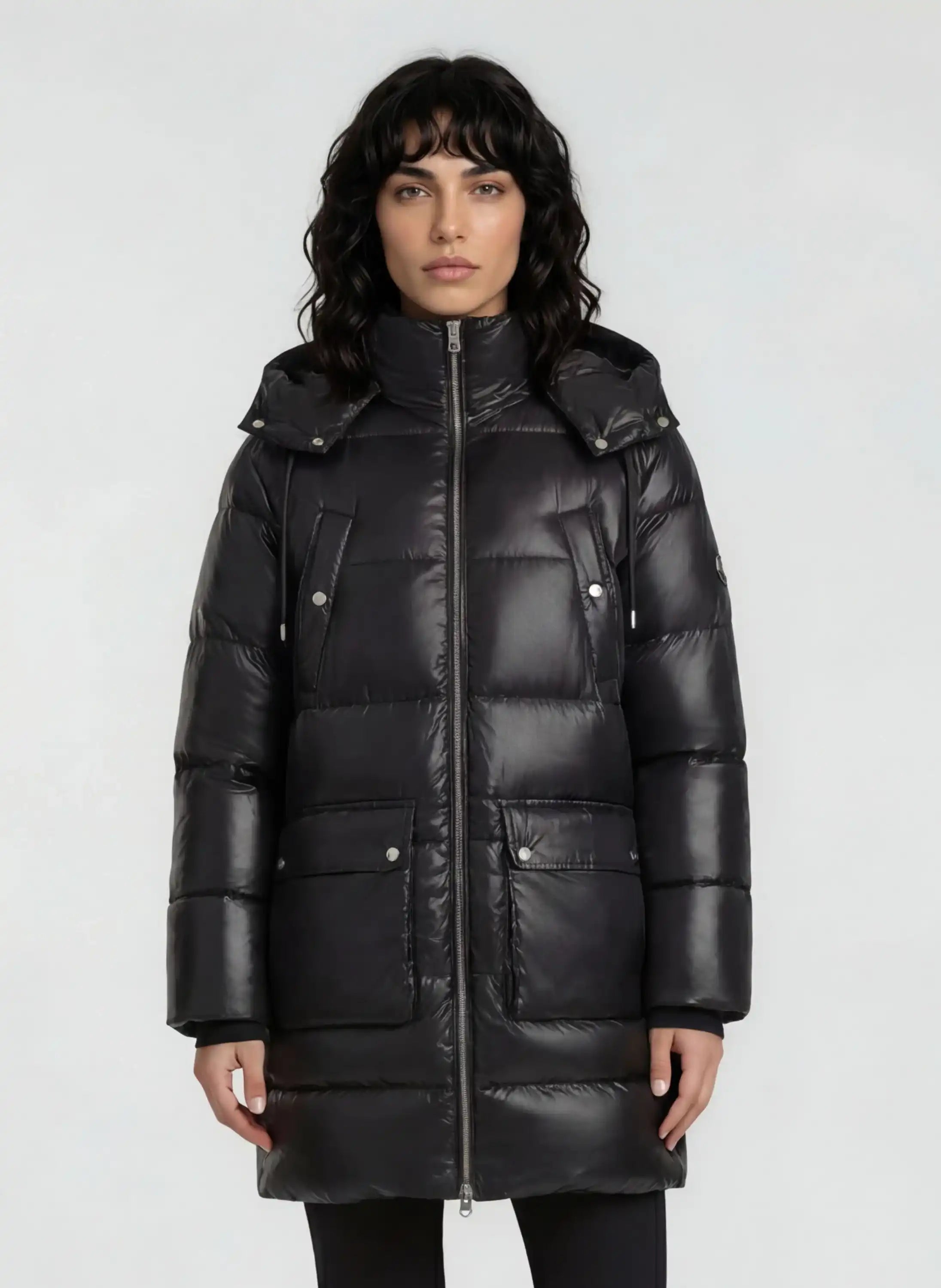 Eine Person mit schulterlangem, gewelltem, dunklem Haar trägt die trueprodigy Damen Winterjacke mit abnehmbarer Kapuze im Regular Fit, mit glänzendem Steppmuster, hohem Kragen, Reißverschluss und zwei Vordertaschen vor hellem Hintergrund.
