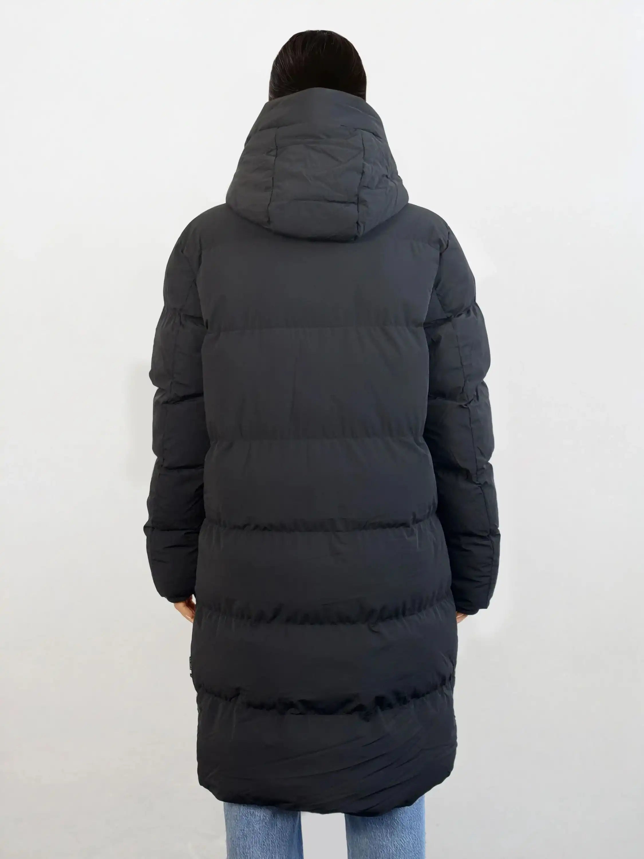 Man sieht eine Person von hinten, die den trueprodigy Damen Parka mit fester Kapuze im Regular Fit in Schwarz mit horizontaler Steppung trägt. Die wasserabweisende Jacke wird mit einer hellblauen Jeans vor einem schlichten, hellen Hintergrund kombiniert.
