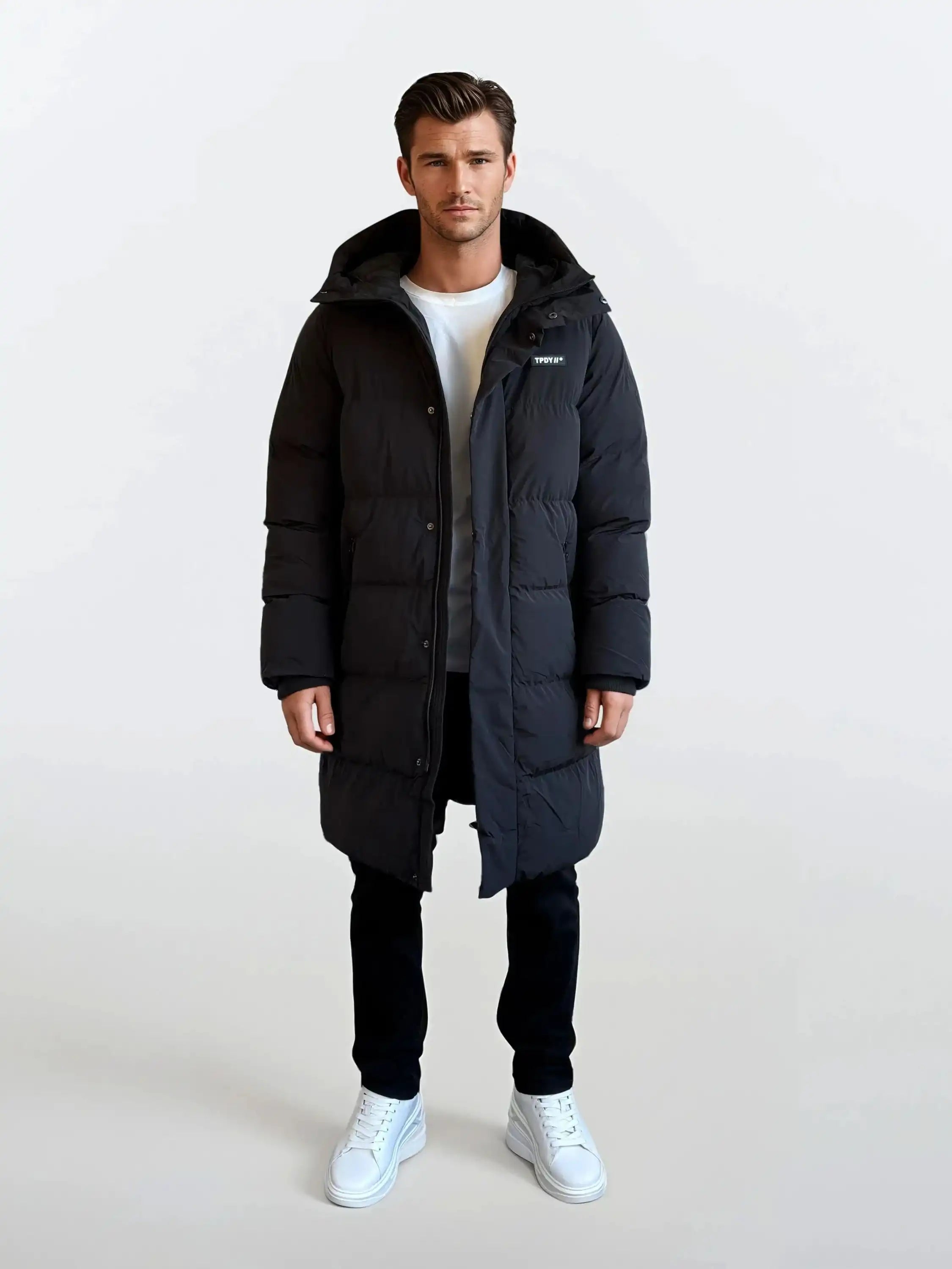 Ein Mann sieht nach vorne und trägt einen trueprodigy Herren Parka mit fester Kapuze im Regular Fit - lang, schwarz und wasserabweisend - über einem weißen T-Shirt, einer schwarzen Hose und weißen Turnschuhen. Der Hintergrund ist einfarbig weiß.