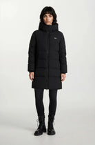 Eine Person steht mit dem Gesicht nach vorne und trägt einen trueprodigy Damen Parka mit fester Kapuze im Regular Fit, eine schwarze Hose und schwarze Schnürstiefel vor einem schlichten hellen Hintergrund.