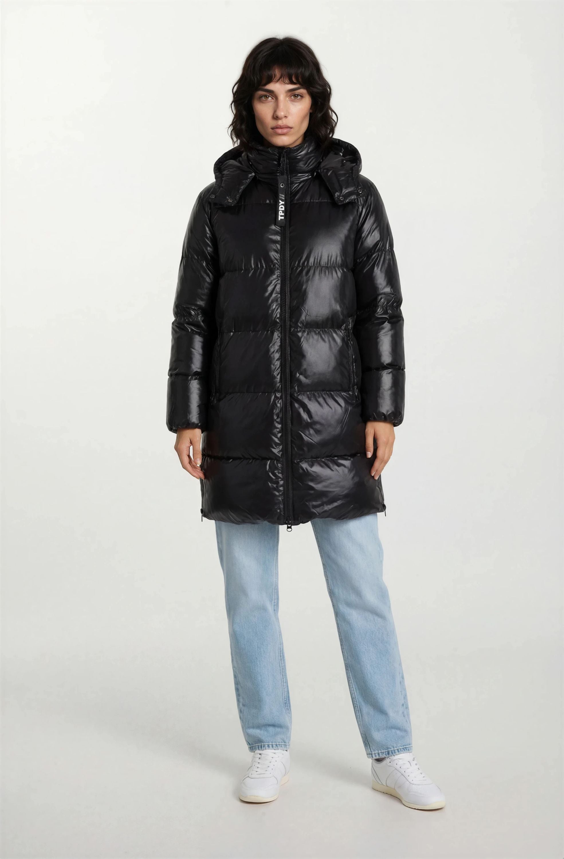 Eine Person blickt nach vorne und trägt eine trueprodigy Damen Winterjacke mit hochglänzender Oberfläche im Regular Fit, hellblaue Jeans und weiße Turnschuhe vor einem schlichten hellen Hintergrund.