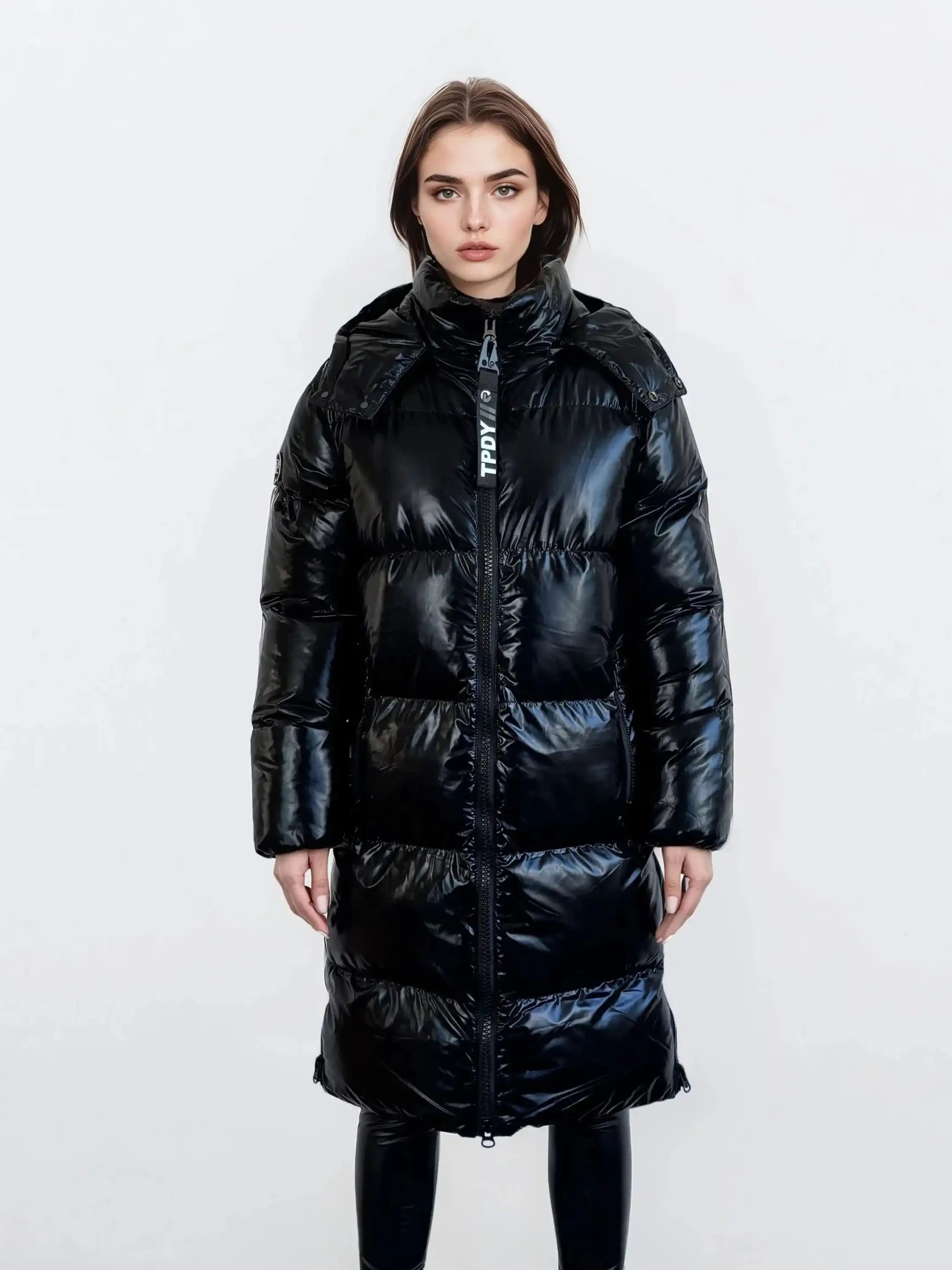 Eine Frau mit glattem braunem Haar und neutralem Gesichtsausdruck modelliert die trueprodigy Damen Winterjacke mit hochglänzender Oberfläche im Regular Fit, vorne mit Reißverschluss und abnehmbarer Kapuze, über einer schwarzen Hose vor einem schlichten weißen Hintergrund.