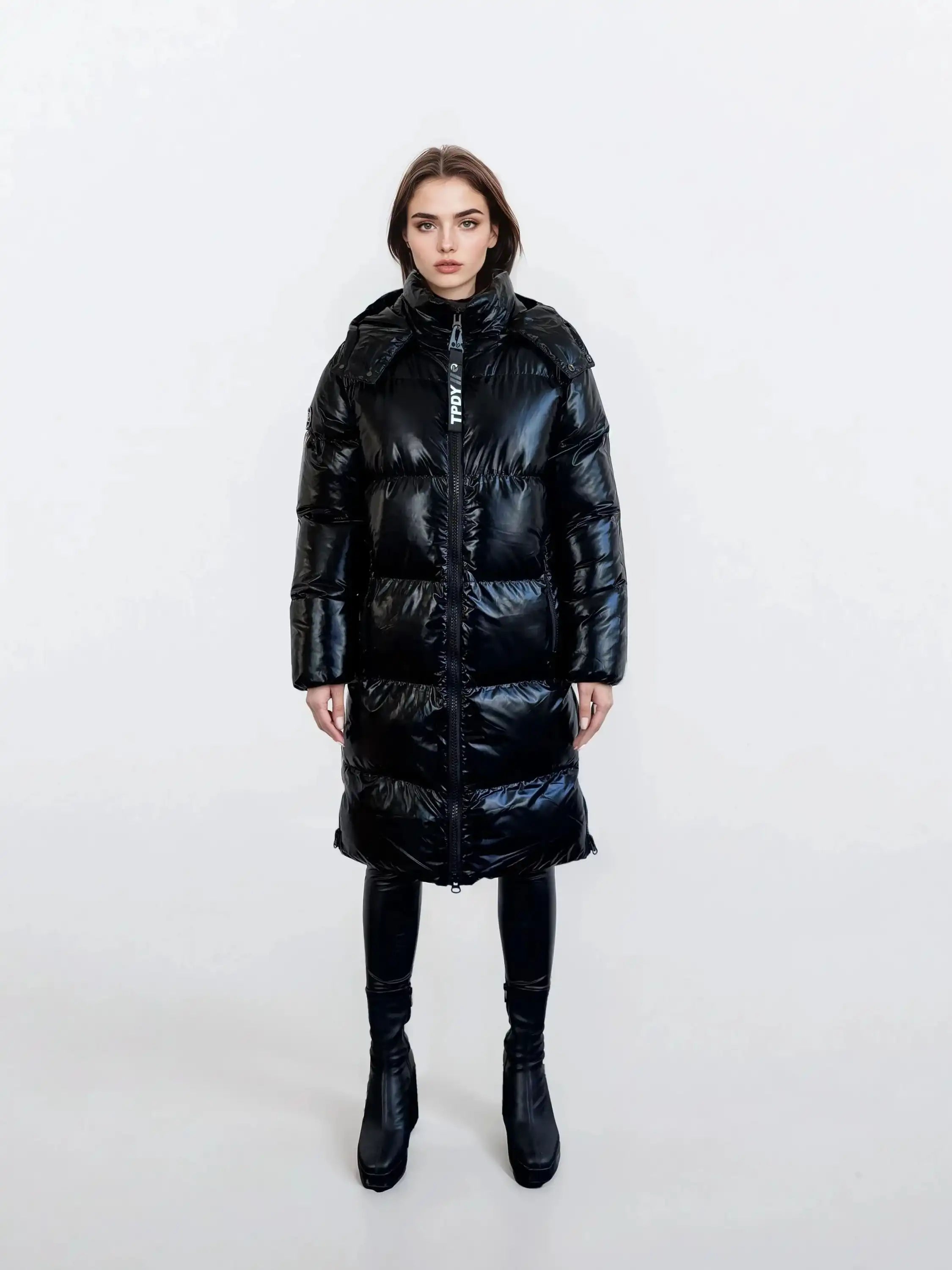 Eine Person steht mit dem Gesicht nach vorne vor einem schlichten weißen Hintergrund und trägt die trueprodigy Damen Winterjacke mit hochglänzender Oberfläche im Regular Fit, gepaart mit einer schwarzen Hose und schwarzen Ankle Boots.