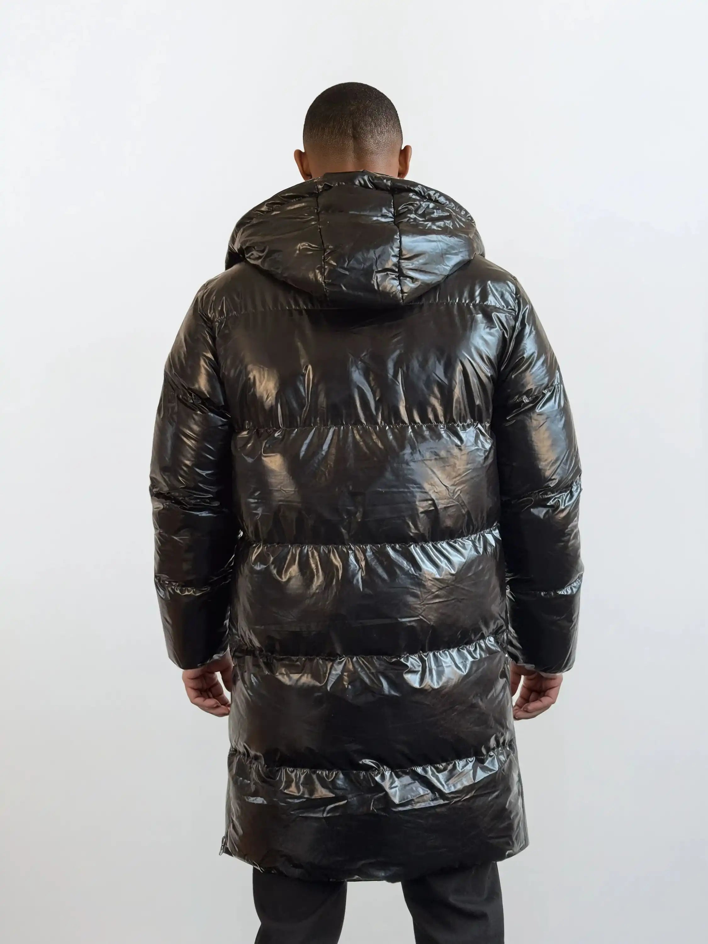 Eine Person blickt von der Kamera weg und trägt die trueprodigy Herren Winterjacke mit hochglänzender Oberfläche im Regular Fit. Die lange schwarze Pufferjacke mit Kapuze hebt sich von dem hellen, schlichten Hintergrund ab.