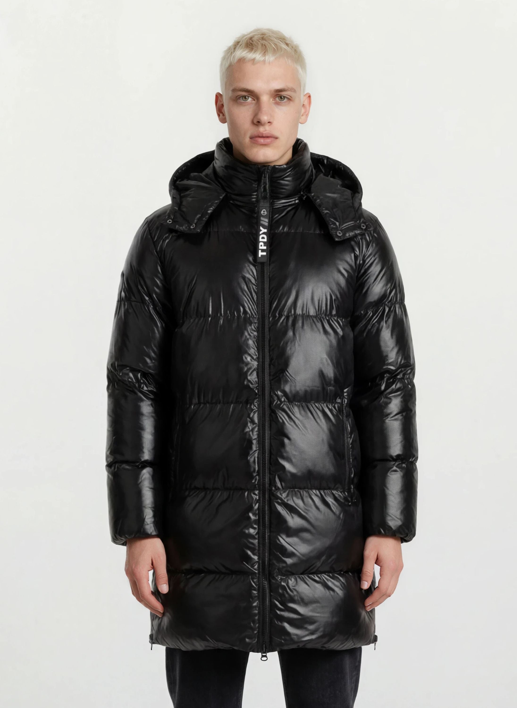 Eine Person blickt vor einem hellen Hintergrund nach vorne und trägt die trueprodigy Herren Winterjacke mit hochglänzender Oberfläche im Regular Fit, die mit einem hohen Kragen, einer abnehmbaren Kapuze und einem Frontreißverschluss für einen eleganten Look ausgestattet ist.
