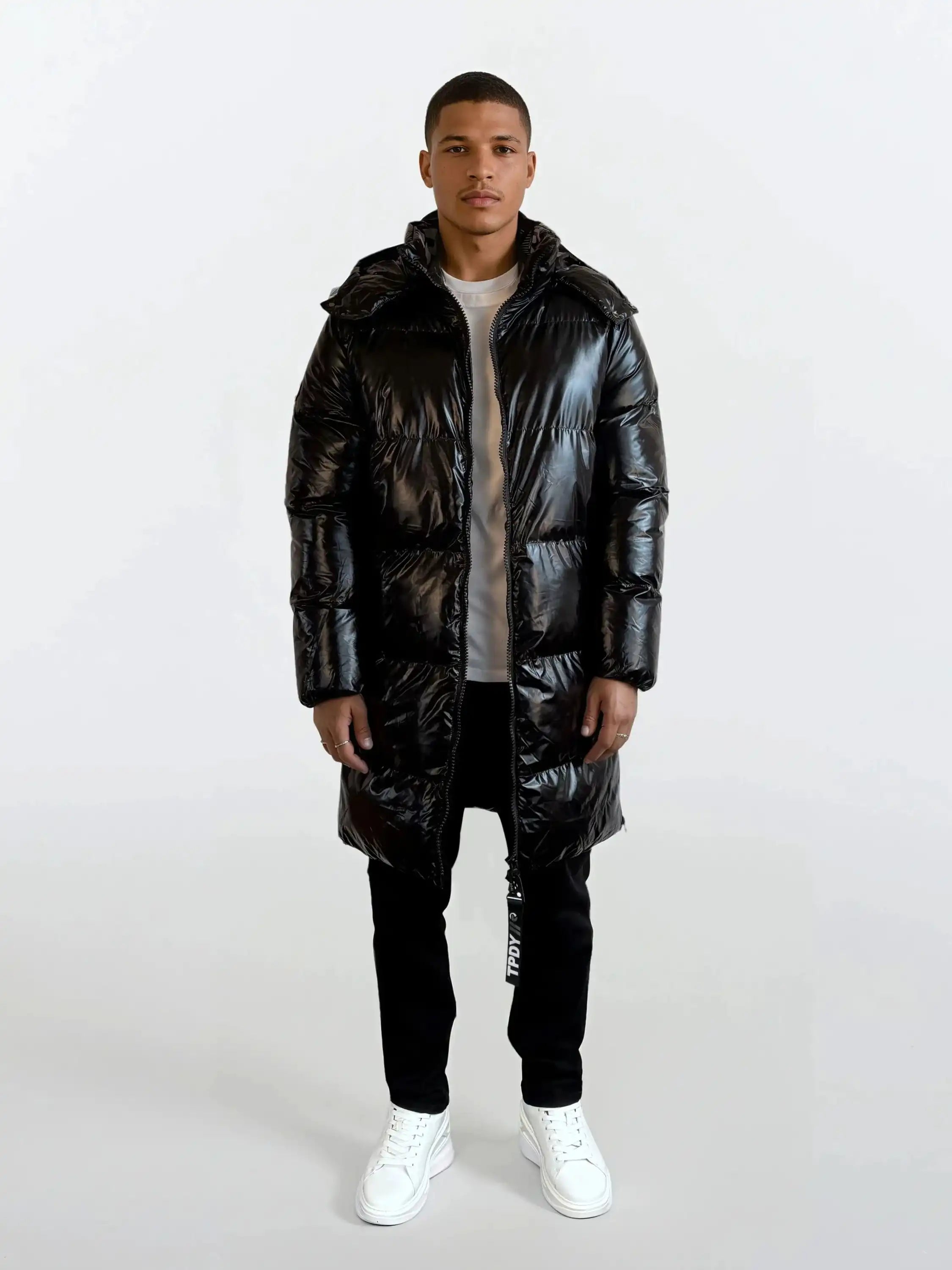 Eine Person steht mit Blick nach vorne in einer glänzenden schwarzen trueprodigy Herren Winterjacke mit hochglänzender Oberfläche im Regular Fit mit abnehmbarer Kapuze, schwarzer Hose, hellem Hemd und weißen Turnschuhen vor einem schlichten weißen Hintergrund.