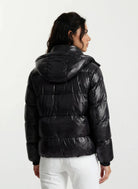 Eine Person mit langen dunklen Haaren, von hinten gesehen, trägt eine trueprodigy Damen Winterjacke mit hochglänzender Oberfläche im Regular Fit und eine weiße Hose und steht vor einem schlichten hellen Hintergrund.