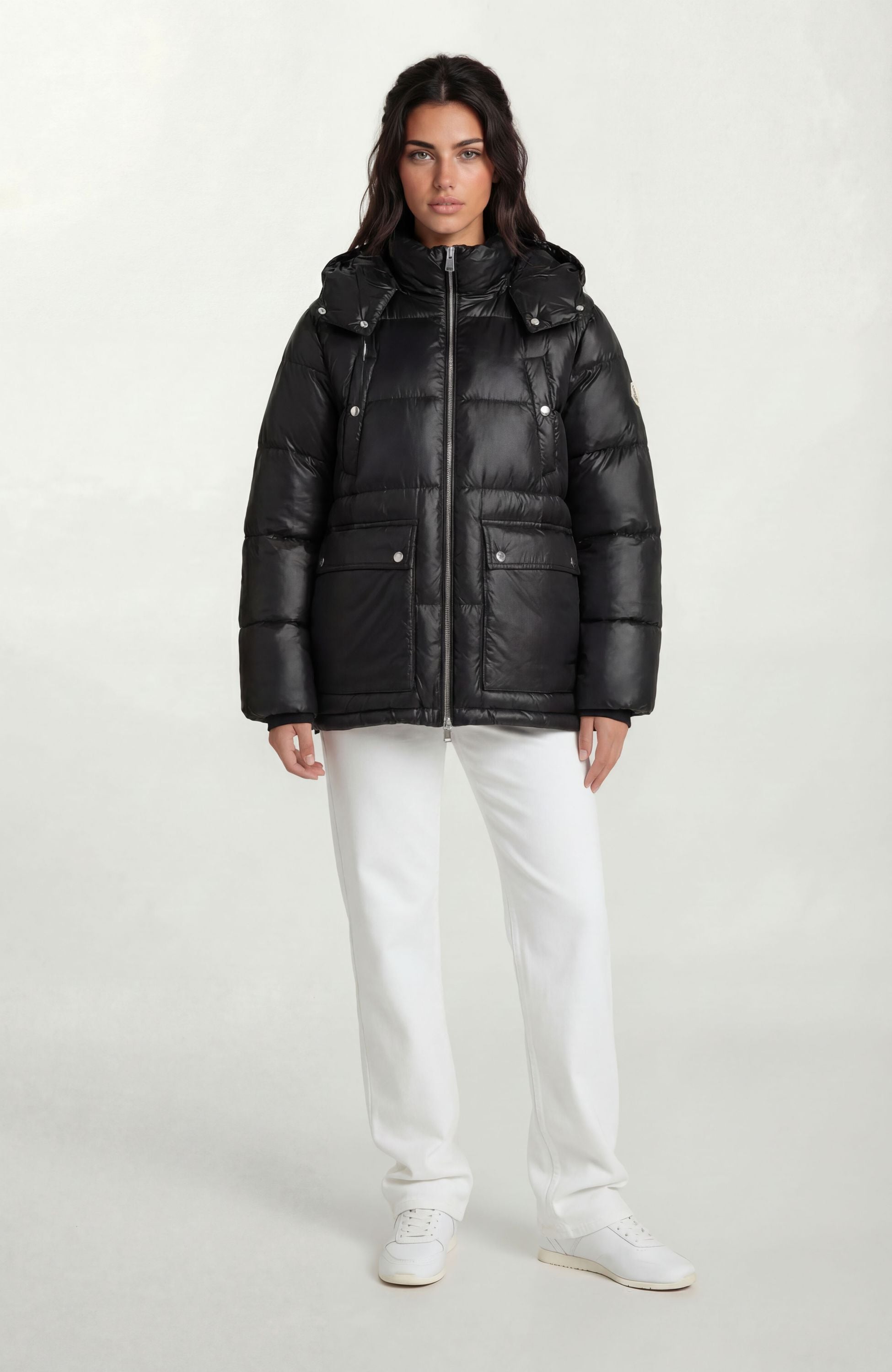 Eine Person steht mit dem Gesicht nach vorne und trägt eine trueprodigy Damen Winterjacke mit hochglänzender Oberfläche im Regular Fit, eine weiße Hose und weiße Turnschuhe vor einem hellgrauen Hintergrund.