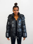 Eine Frau mit dunklem Haar in einem Haarknoten trägt die trueprodigy Damen Winterjacke mit hochglänzender Oberfläche im Regular Fit über einem schwarzen Oberteil und einer Hose vor einem schlichten weißen Hintergrund.