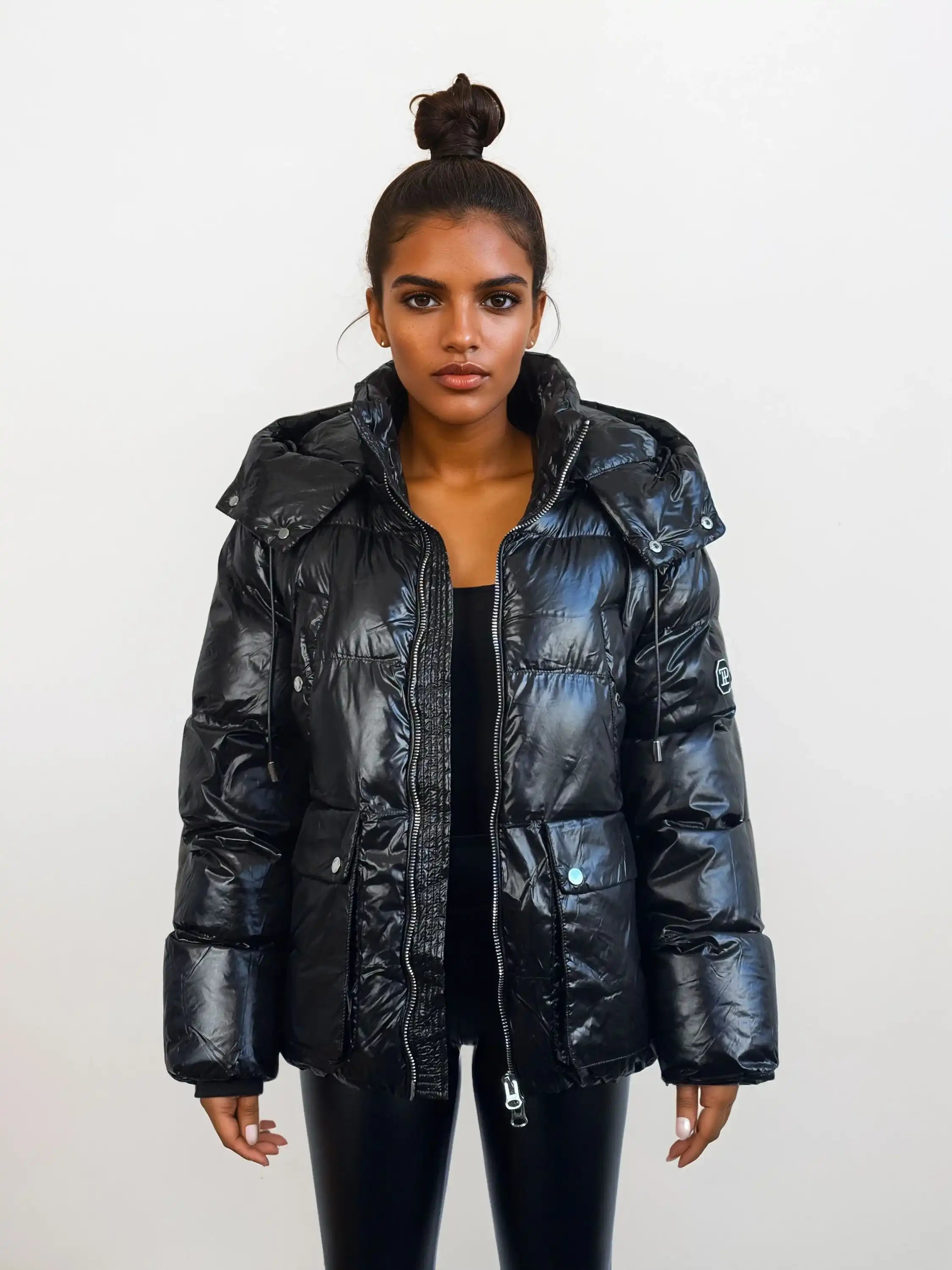Eine Frau mit dunklem Haar in einem Haarknoten trägt die trueprodigy Damen Winterjacke mit hochglänzender Oberfläche im Regular Fit über einem schwarzen Oberteil und einer Hose vor einem schlichten weißen Hintergrund.