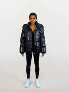 Eine Frau blickt nach vorne in einer trueprodigy Damen Winterjacke mit hochglänzender Oberfläche im Regular Fit, gepaart mit schwarzen Leggings und weißen Sneakers. Sie posiert mit entspannten Händen an den Seiten vor einem hellen, schlichten Hintergrund.
