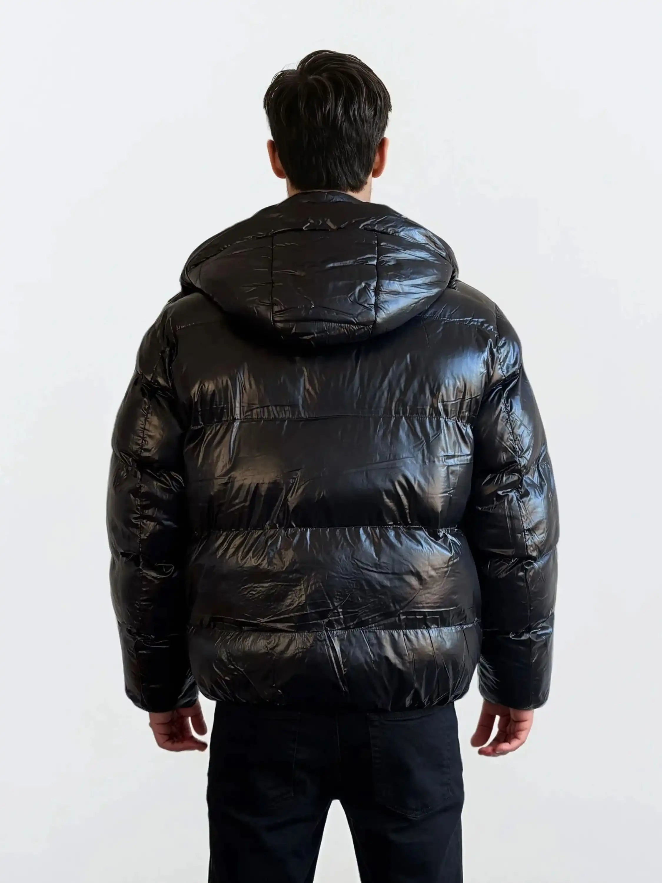 Eine Person steht in einer schwarzen Hose und einer trueprodigy Herren Winterjacke mit hochglänzender Oberfläche im Regular Fit mit Kapuze vor einem schlichten hellen Hintergrund vor der Kamera.