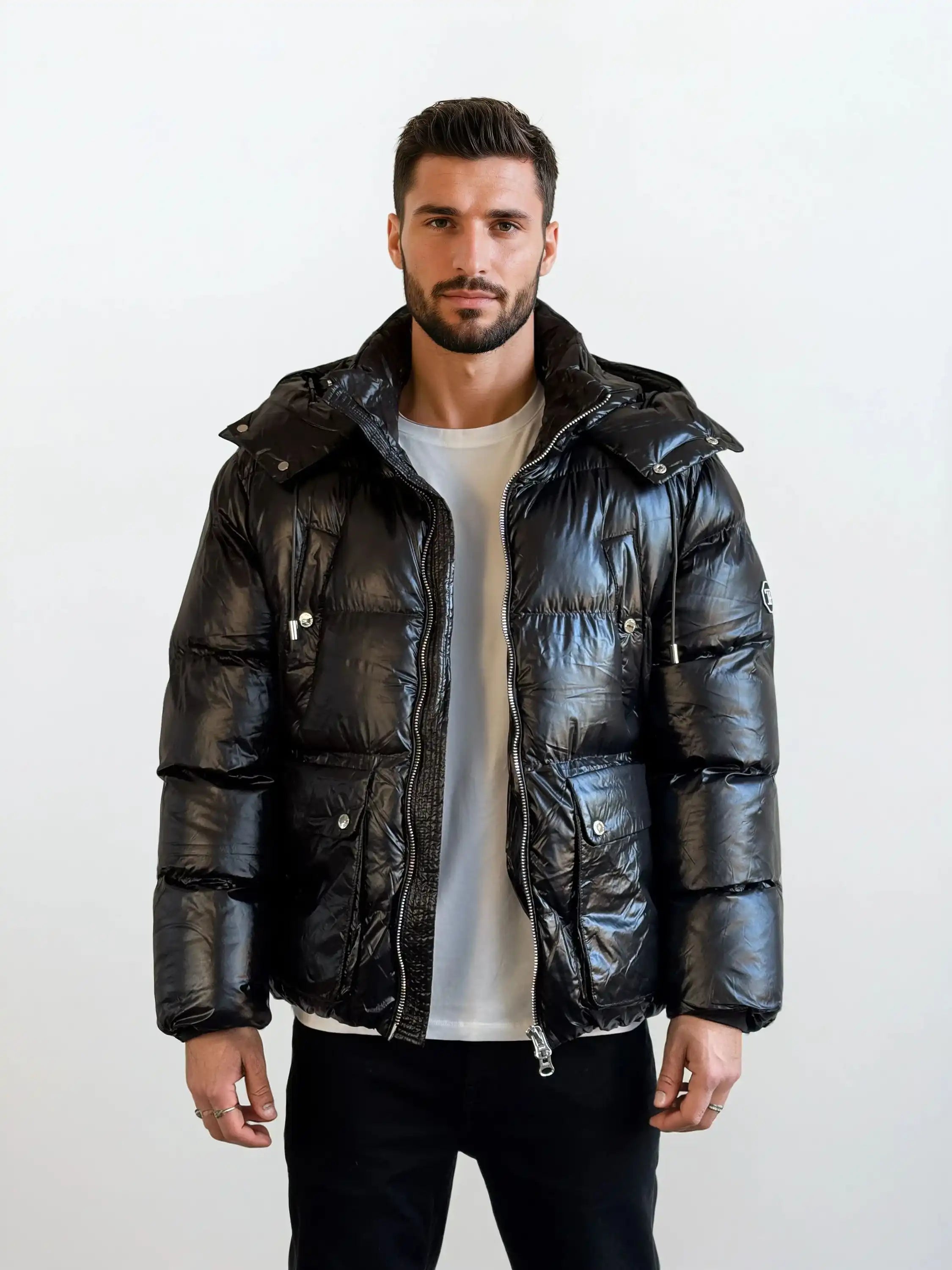 Ein Mann mit kurzen dunklen Haaren und einem Bart steht mit dem Gesicht nach vorne und trägt die trueprodigy Herren Winterjacke mit hochglänzender Oberfläche im Regular Fit über einem weißen T-Shirt und einer schwarzen Hose vor einem schlichten hellen Hintergrund.