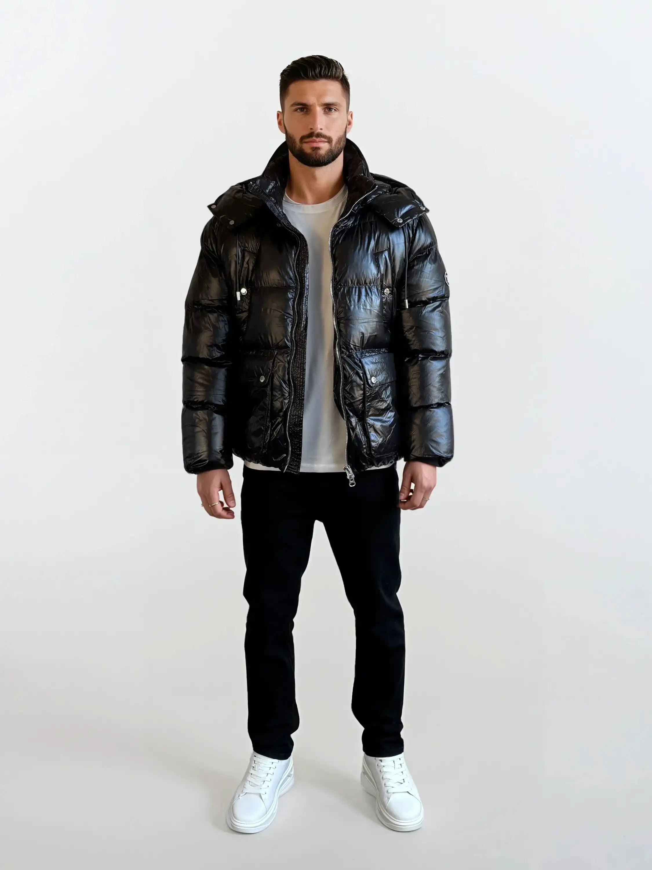 Ein Mann steht mit dem Gesicht nach vorne vor einem schlichten weißen Hintergrund und trägt die trueprodigy Herren Winterjacke mit hochglänzender Oberfläche im Regular Fit, gepaart mit einem weißen T-Shirt, einer schwarzen Hose und weißen Sneakers.