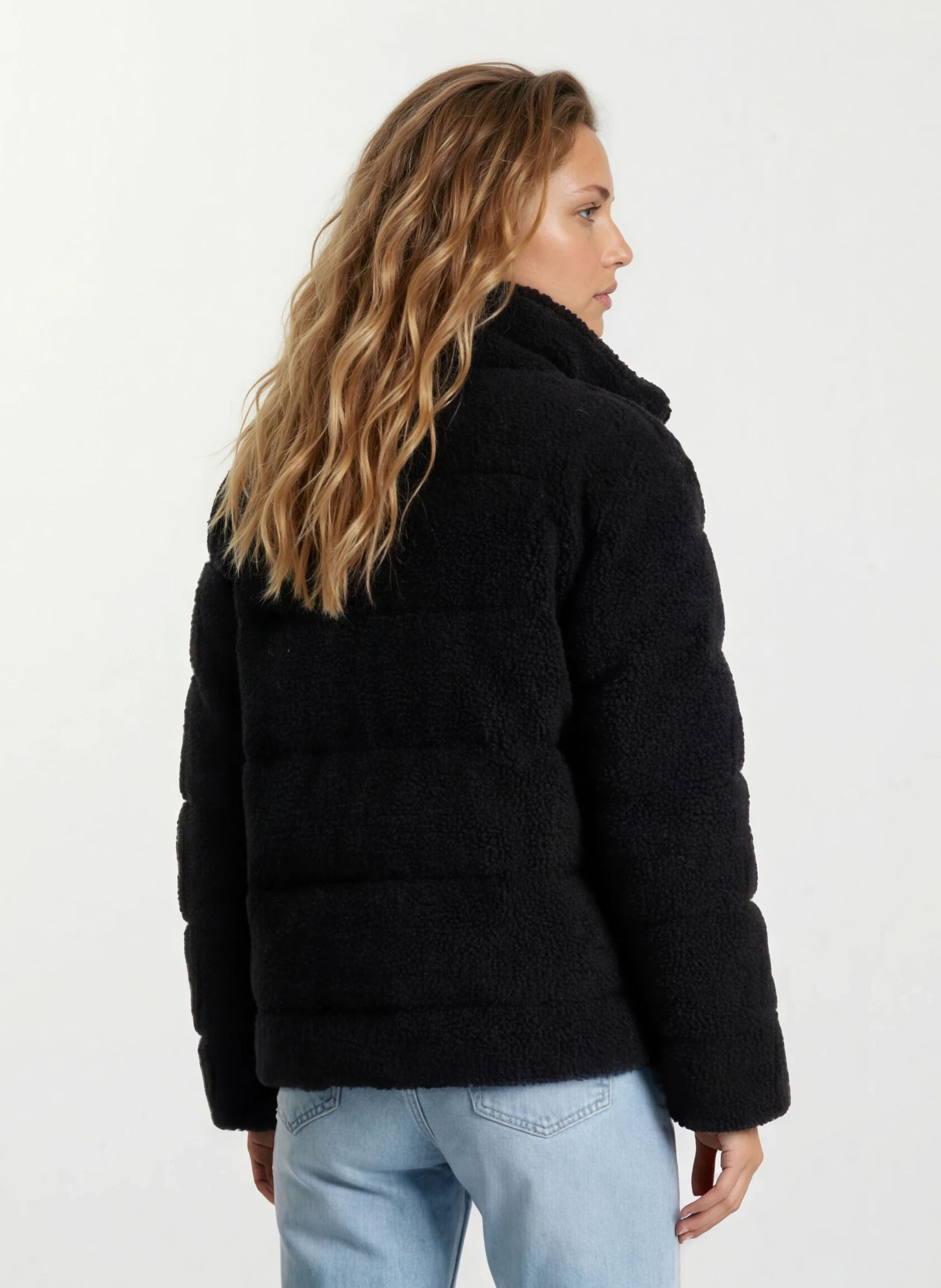 Eine Person mit langen, gewellten blonden Haaren wird von hinten gezeigt, die eine trueprodigy Damen Winterjacke mit flauschiger Sherpa-Oberfläche im Oversized Fit in Schwarz und hellblauen Jeans vor weißem Hintergrund trägt.