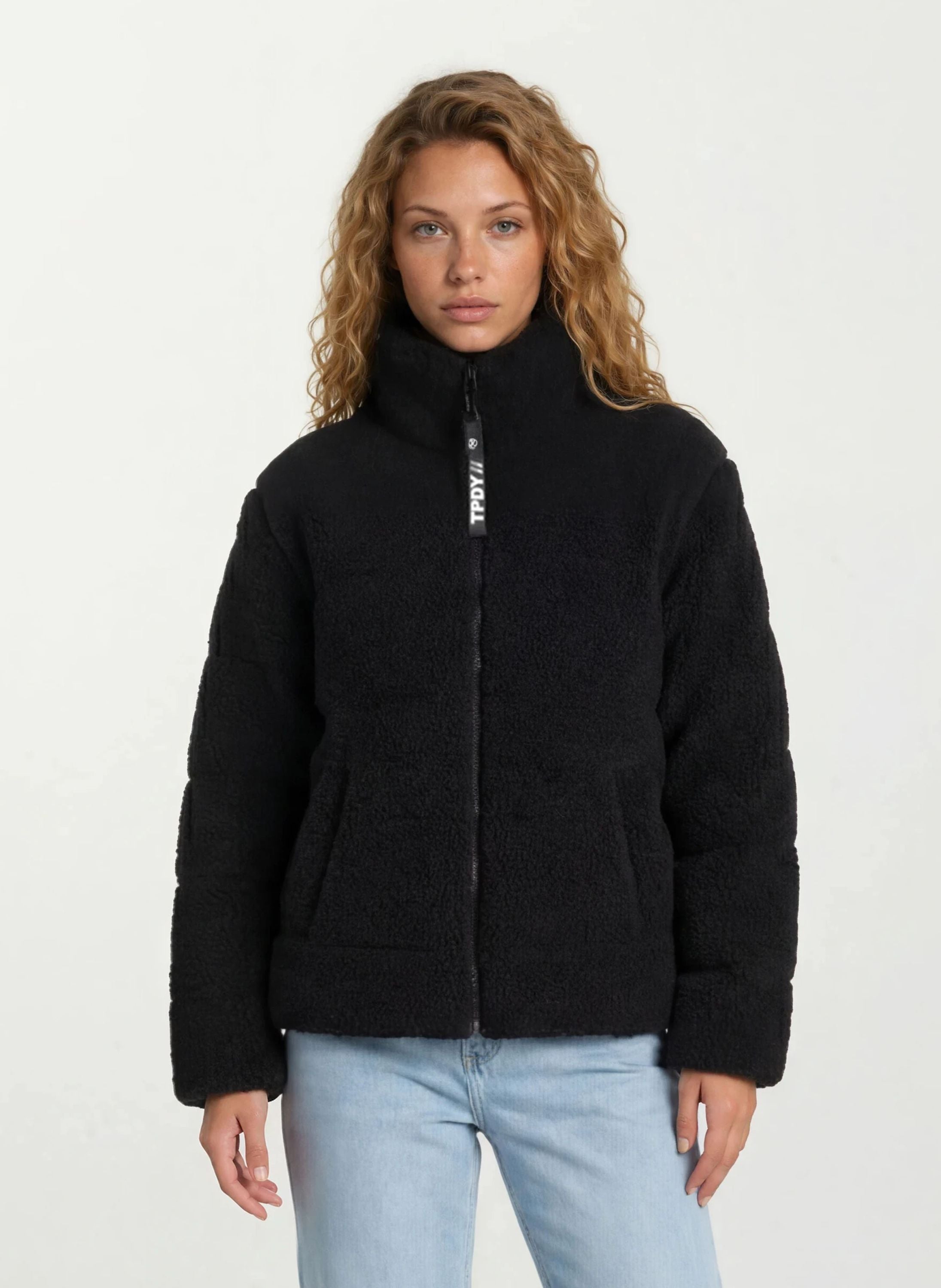 Eine Person mit langen lockigen Haaren trägt die trueprodigy Damen Winterjacke mit flauschiger Sherpa-Oberfläche im Oversized Fit in Schwarz und hellblauen Jeans vor einem schlichten weißen Hintergrund.