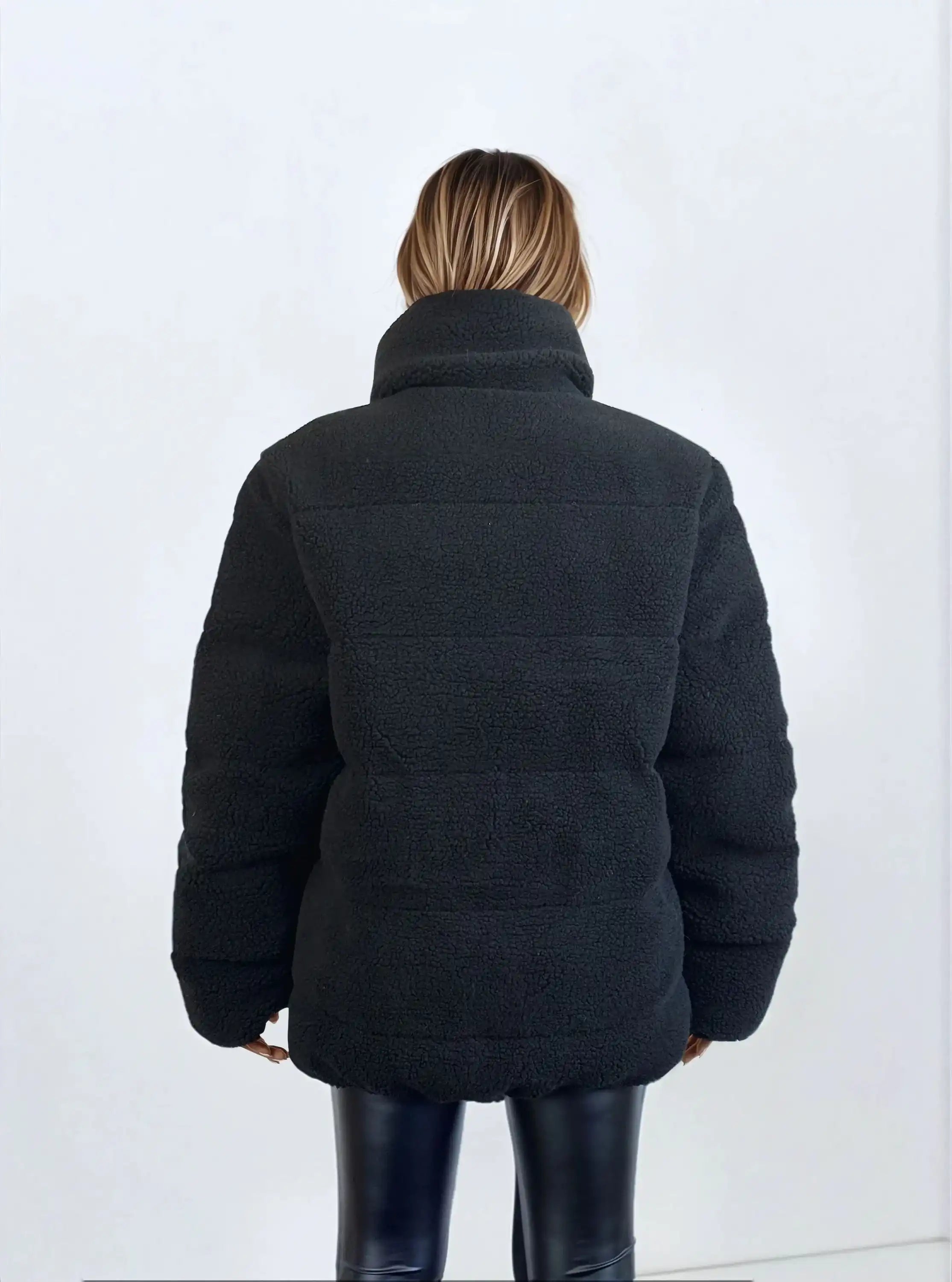 Eine Person mit schulterlangem Haar, von hinten gesehen, trägt eine schwarze Damen Winterjacke mit flauschiger Sherpa-Oberfläche im Oversized Fit von trueprodigy und schwarze Lederhose. Der Hintergrund ist einfarbig und hell.