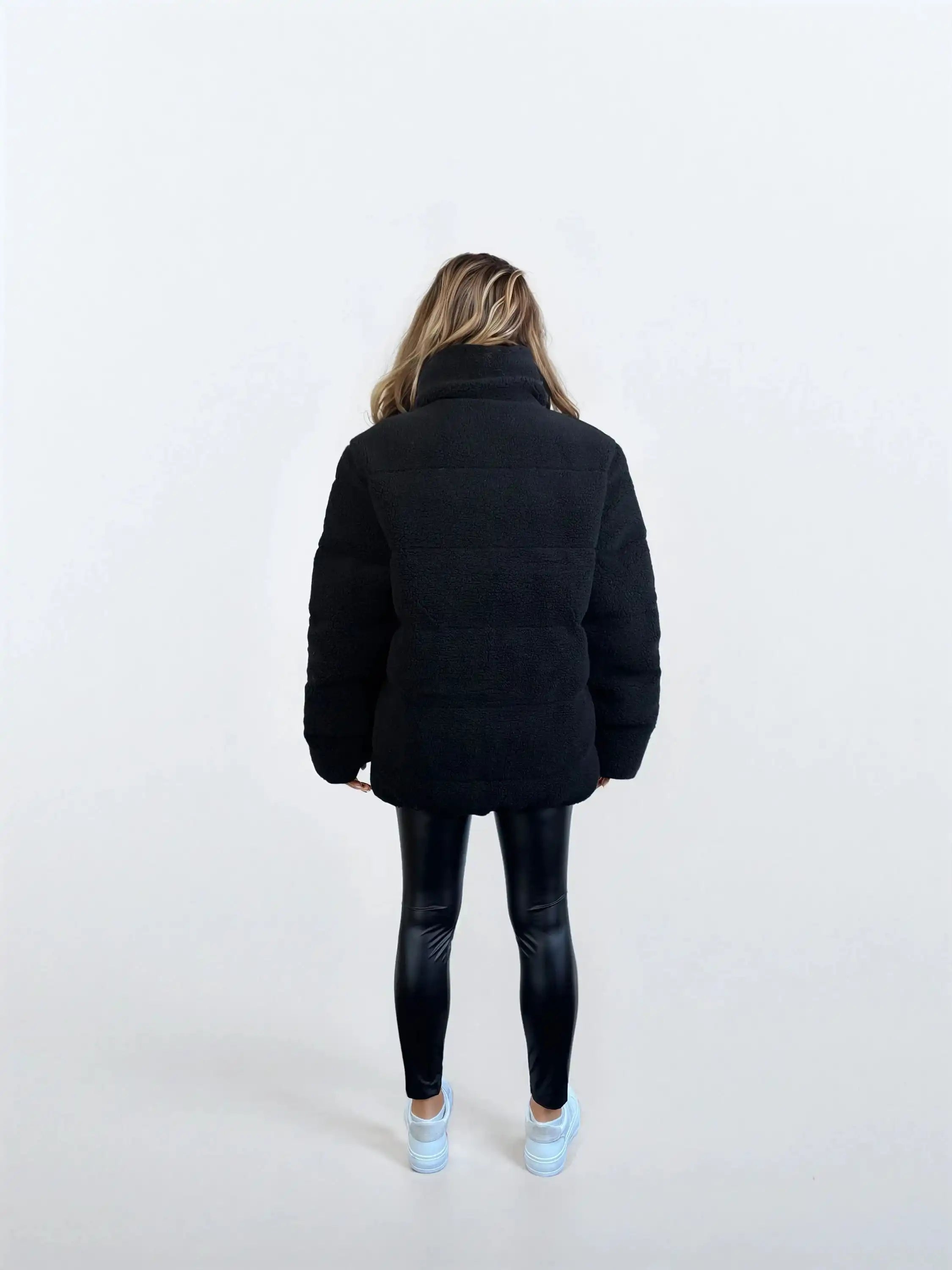 Eine Person mit langen, hellbraunen Haaren steht mit dem Gesicht zur Kamera und trägt die trueprodigy Damen Winterjacke mit flauschiger Sherpa-Oberfläche im Oversized Fit, schwarz glänzende Leggings und weiße Turnschuhe vor einem schlichten weißen Hintergrund.