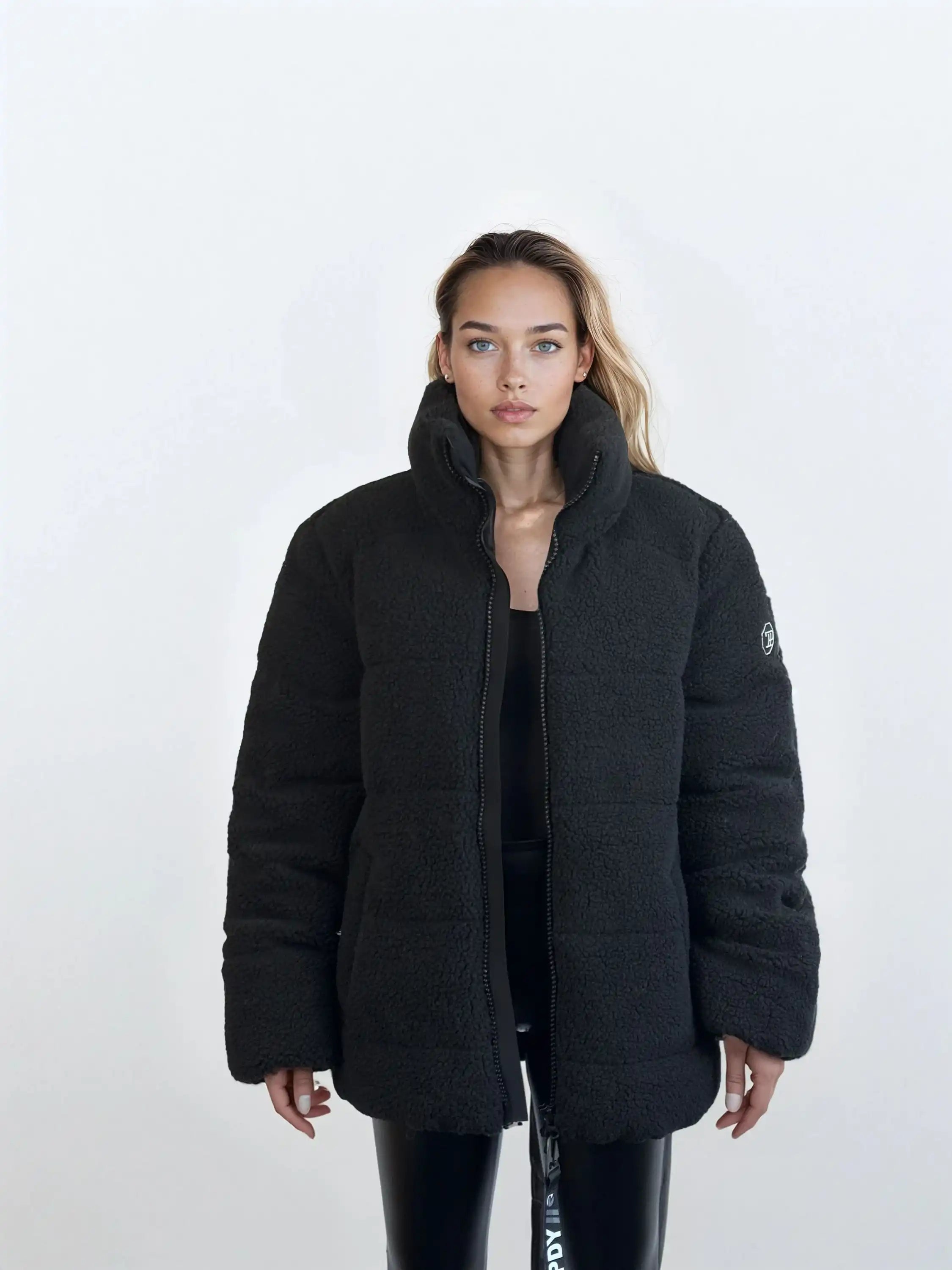 Eine Frau mit langen, hellbraunen Haaren steht vor einem schlichten weißen Hintergrund und trägt die trueprodigy Damen Winterjacke mit flauschiger Sherpa-Oberfläche im Oversized Fit über schwarzer Kleidung. Sie blickt mit neutralem Blick in die Kamera.