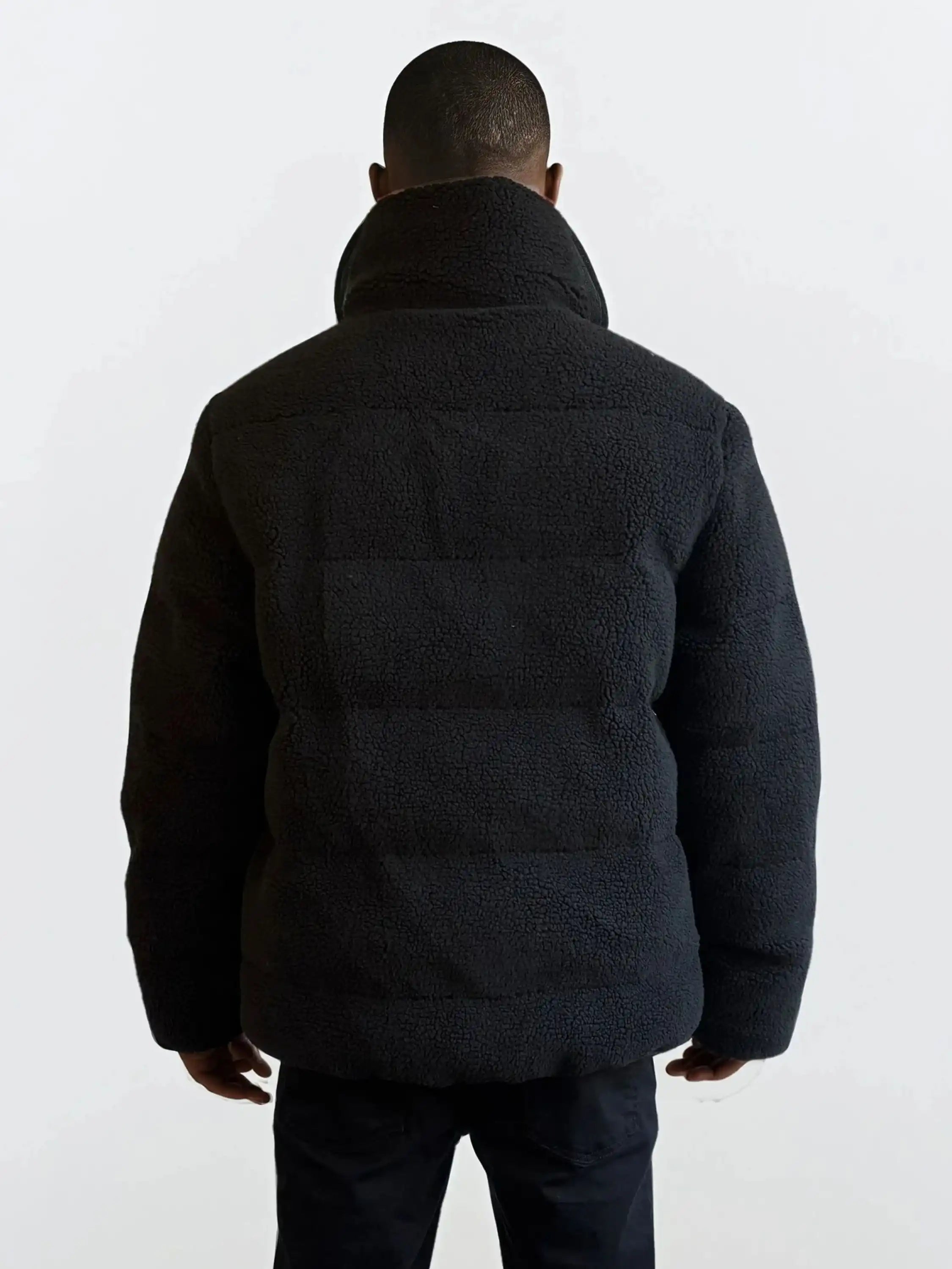 Eine Person steht mit dem Rücken zur Kamera und trägt eine schwarze trueprodigy Herren Winterjacke mit flauschiger Sherpa-Oberfläche im Oversized Fit und eine dunkle Hose vor einem schlichten hellen Hintergrund.
