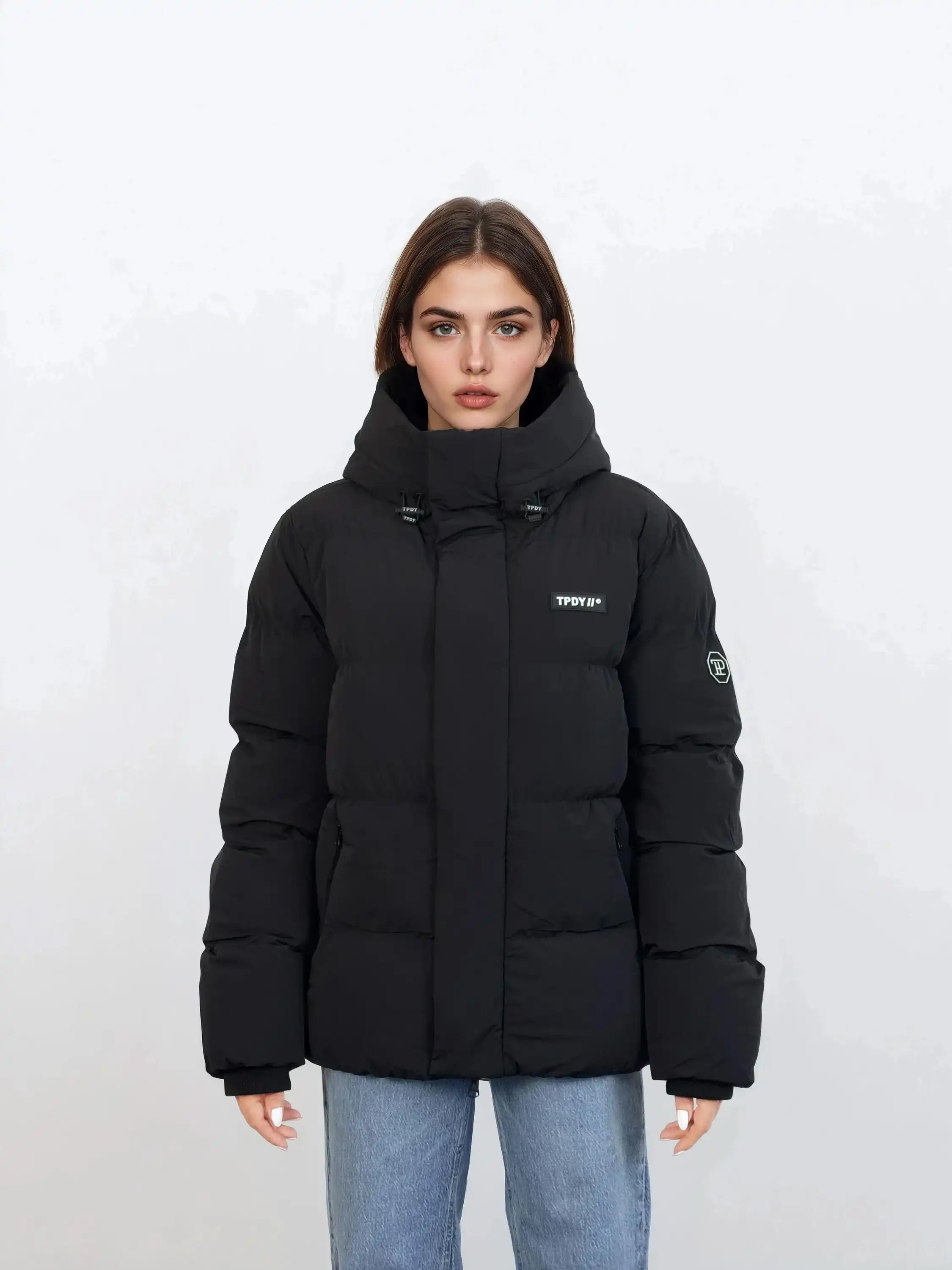 Eine Person mit glattem braunem Haar trägt eine trueprodigy Damen Winterjacke mit fester Kapuze im Oversized Fit in Schwarz mit Logoaufnähern auf der Brust und dem oberen Ärmel, gepaart mit hellblauen Jeans vor einem schlichten weißen Hintergrund.