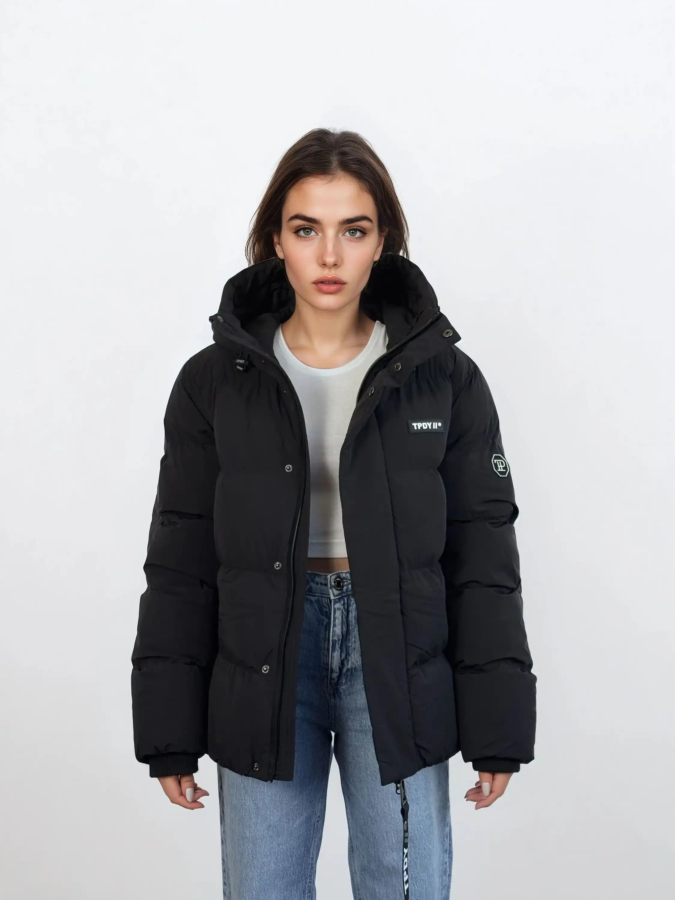 Eine Person steht vor weißem Hintergrund und trägt eine schwarze, wasserabweisende trueprodigy Damen Winterjacke mit fester Kapuze im Oversized Fit über einem hellgrauen Shirt und blauen Jeans. Die Jacke hat einen Aufnäher am Ärmel und den Schriftzug STOFFE auf der Brust.