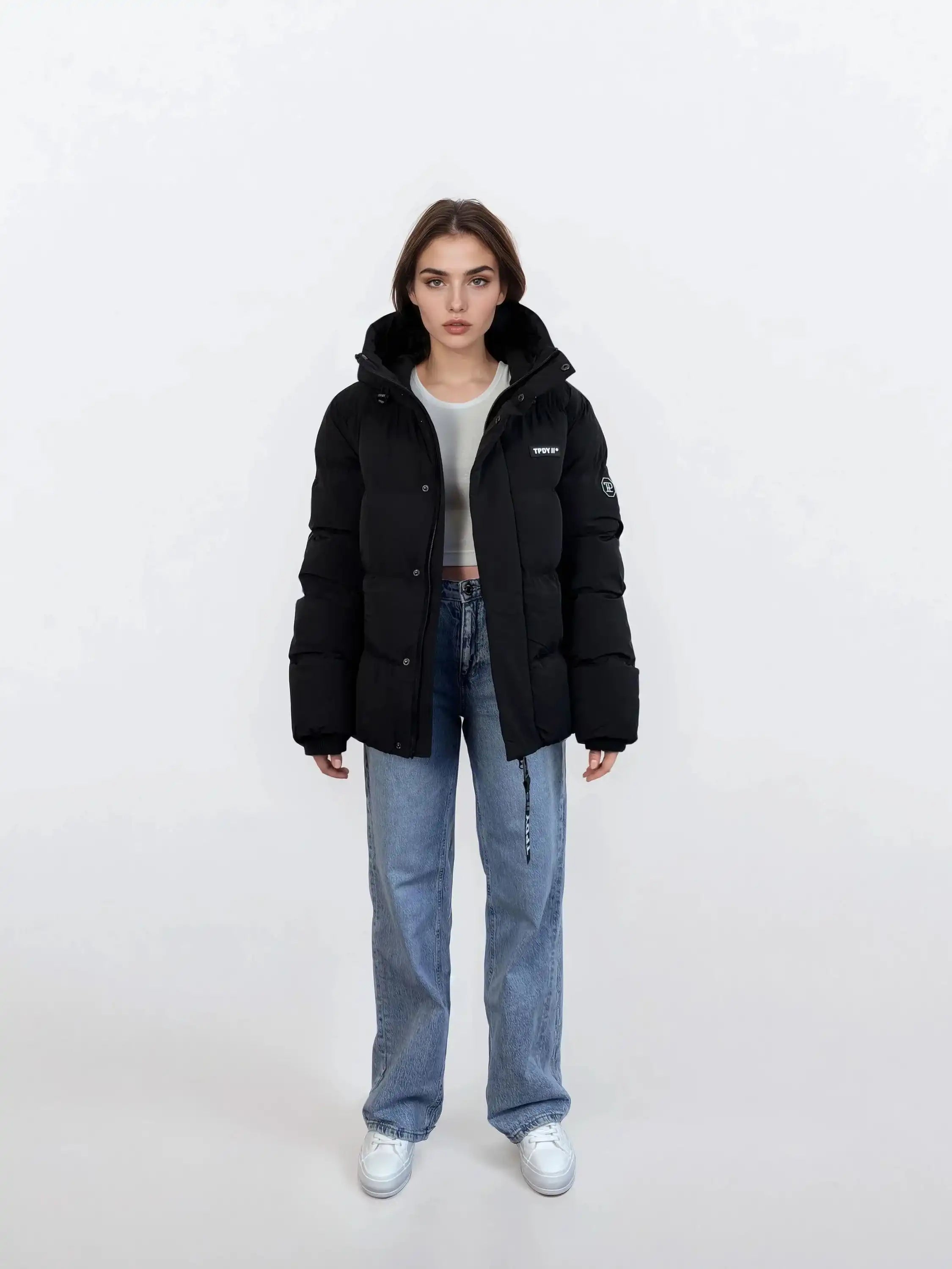 Eine Frau steht vor einem schlichten weißen Hintergrund und trägt eine trueprodigy Damen Winterjacke mit fester Kapuze im Oversized Fit, hellblaue Jeans, ein weißes Kurzarmhemd und weiße Turnschuhe, wobei sie mit den Armen nach vorne schaut.