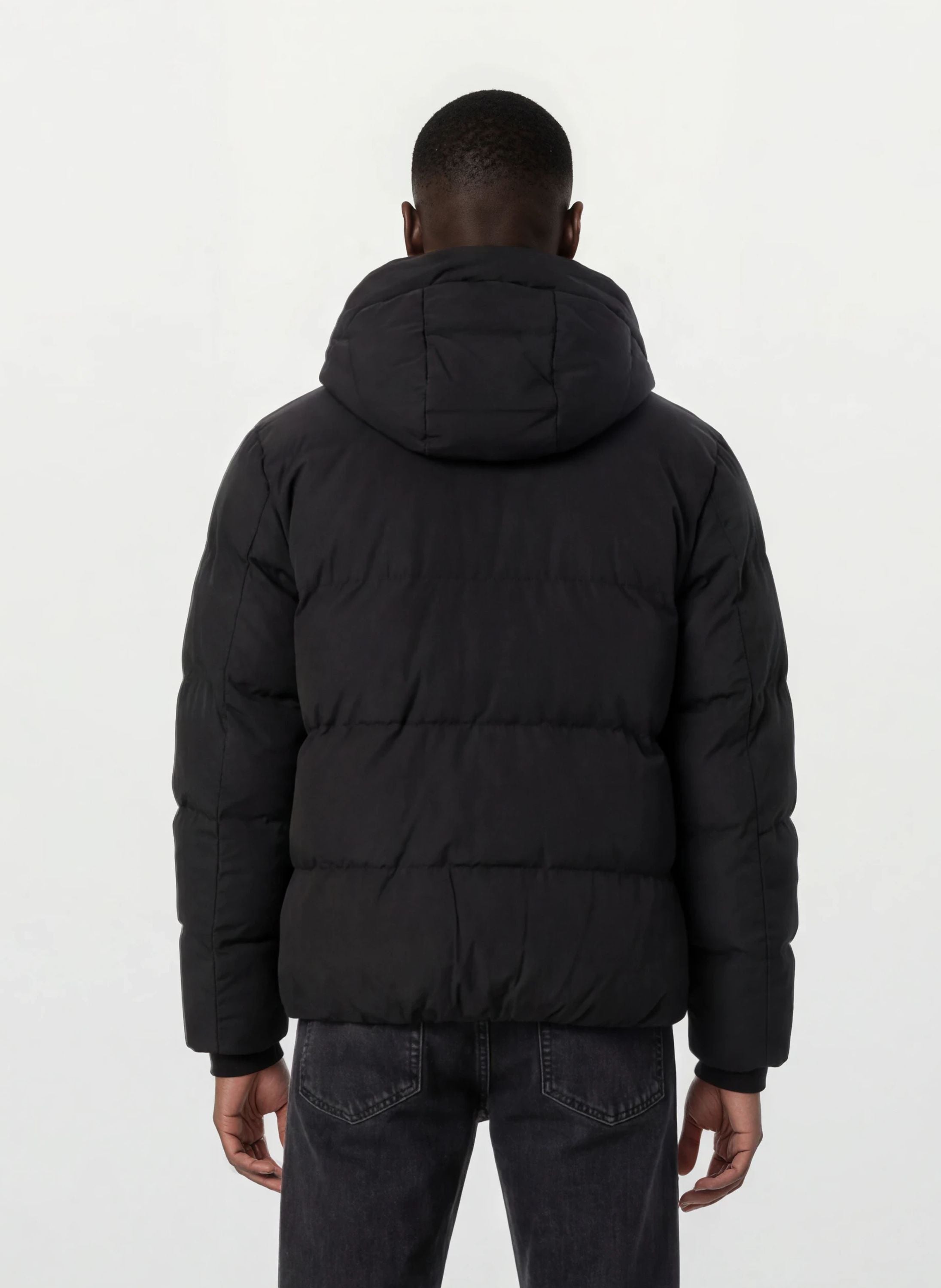 Eine Person mit kurzen Haaren wird von hinten gezeigt, die eine trueprodigy Herren Winterjacke mit fester Kapuze im Oversized Fit und dunkle Jeans trägt. Die Jacke wirkt wasserabweisend, der Hintergrund ist schlicht und hell.