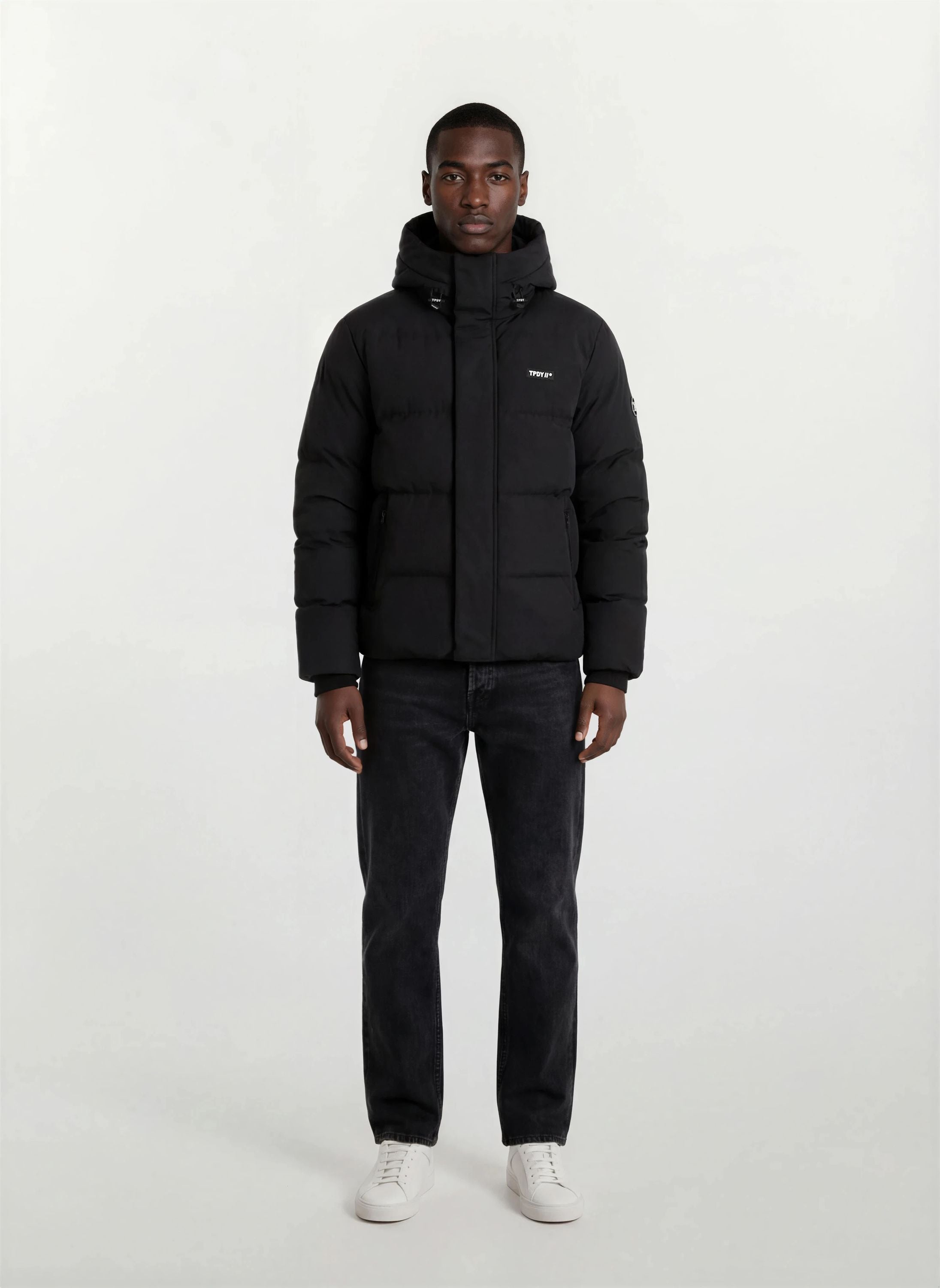 Eine Person steht mit dem Gesicht nach vorne vor einem schlichten weißen Hintergrund und trägt die trueprodigy Herren Winterjacke mit fester Kapuze im Oversized Fit, gepaart mit dunklen Jeans und weißen Sneakers.