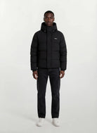 Eine Person steht mit dem Gesicht nach vorne vor einem schlichten weißen Hintergrund und trägt die trueprodigy Herren Winterjacke mit fester Kapuze im Oversized Fit, gepaart mit dunklen Jeans und weißen Sneakers.