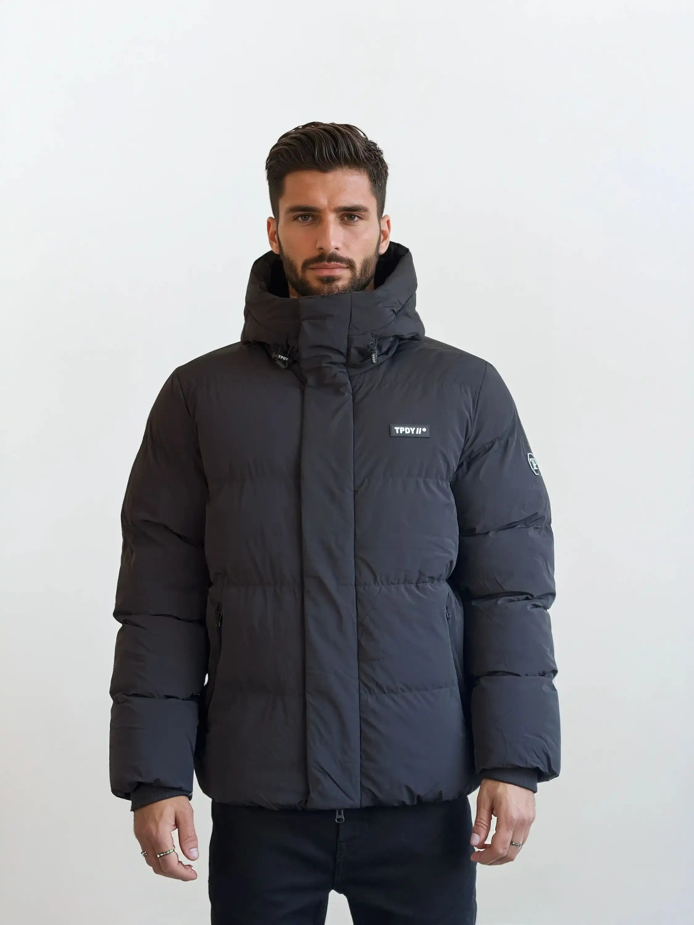 Ein Mann mit kurzem dunklem Haar und Bart trägt eine schwarze trueprodigy Herren Winterjacke mit fester Kapuze im Oversized Fit, wasserabweisend, mit kleinem Logo auf der Brust und Patch am linken Ärmel. Weißer Hintergrund.