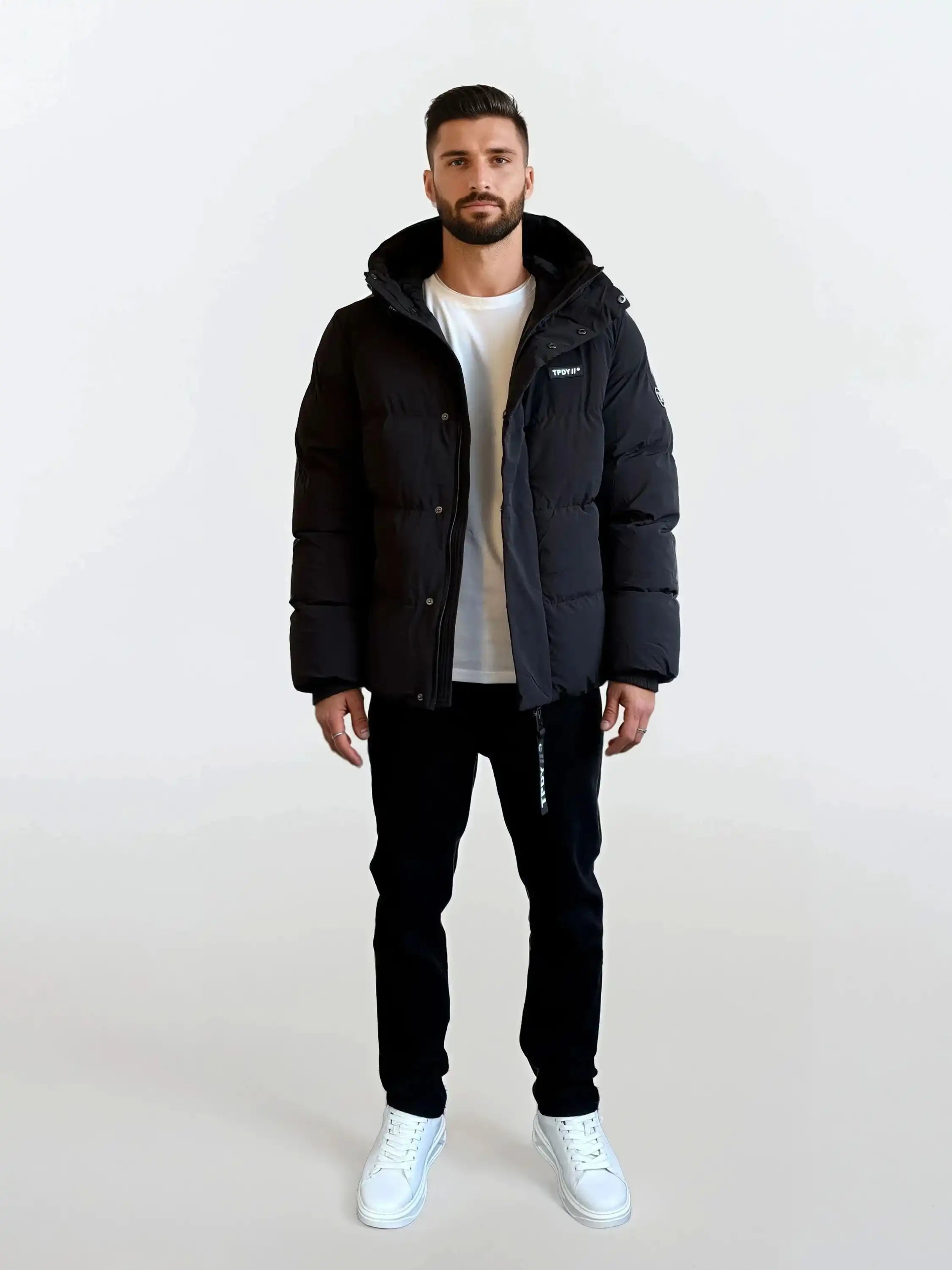Ein Mann steht mit dem Gesicht nach vorne vor einem schlichten weißen Hintergrund und trägt die trueprodigy Herren Winterjacke mit fester Kapuze im Oversized Fit, ein weißes T-Shirt, eine schwarze Hose und weiße Turnschuhe.