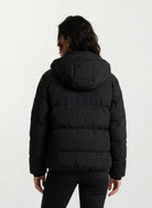 Eine Person steht mit dem Rücken zur Kamera und trägt die trueprodigy Damen Winterjacke mit fester Kapuze im Oversized Fit und schwarzer Hose vor einem einfachen hellen Hintergrund.