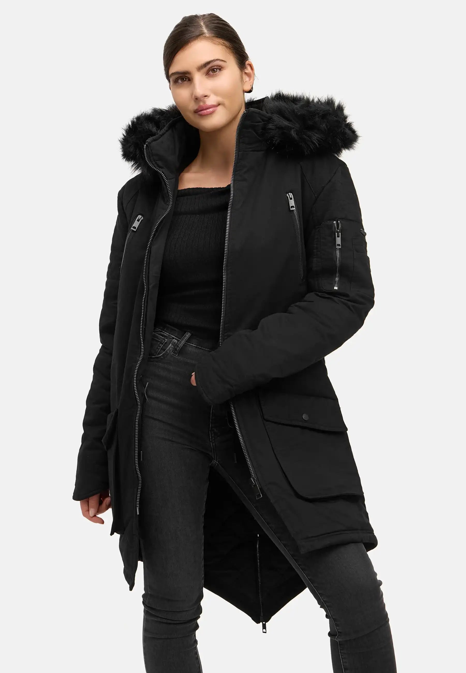 Eine Frau trägt den trueprodigy Harvey F Damenparka mit Kapuze und Kunstfell in Schwarz, gepaart mit einem schwarzen Pullover und einer schmal geschnittenen Jeans vor einem schlichten hellen Hintergrund. Sie steht mit einer Hand in der Tasche und zeigt einen neutralen Gesichtsausdruck.