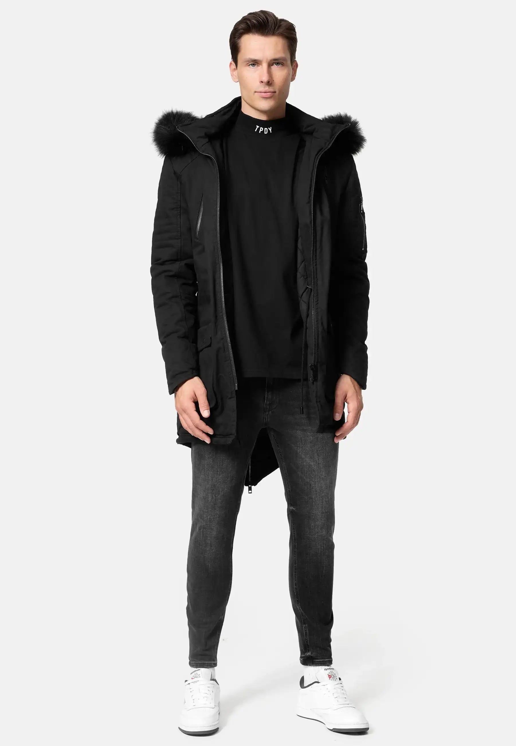 Ein Mann steht mit dem Blick nach vorne und trägt den trueprodigy Harvey Parka mit Kapuze und Kunstfell, gepaart mit einem schwarzen Rollkragenpullover, Slim-Fit-Jeans und weißen Nike-Sneakers vor einem schlichten hellgrauen Hintergrund.
