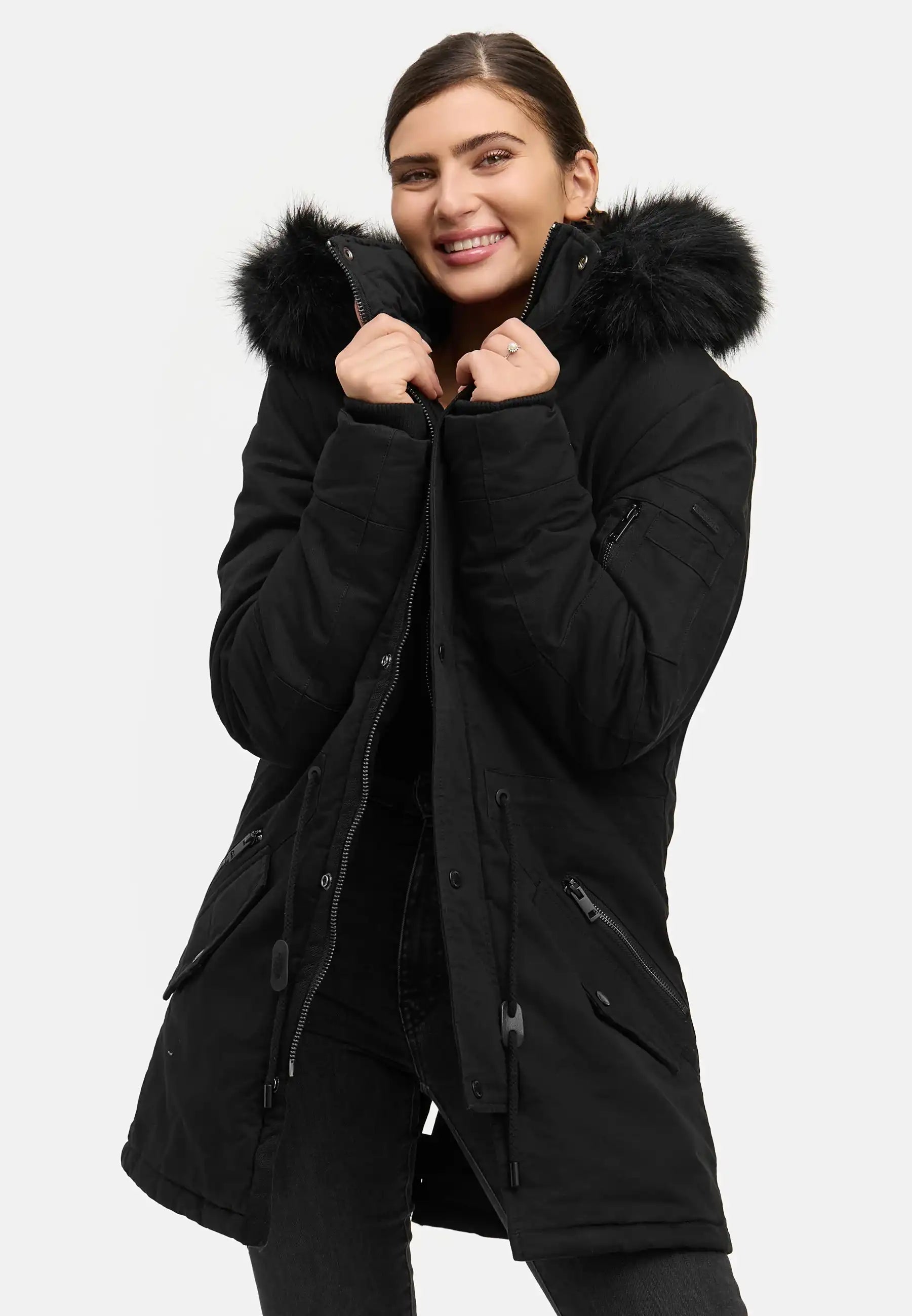 Eine Person lächelt, während sie den trueprodigy Julius F Parka mit Kapuze und Kunstfell in Schwarz trägt und den Kragen mit beiden Händen vor einem schlichten weißen Hintergrund hält.