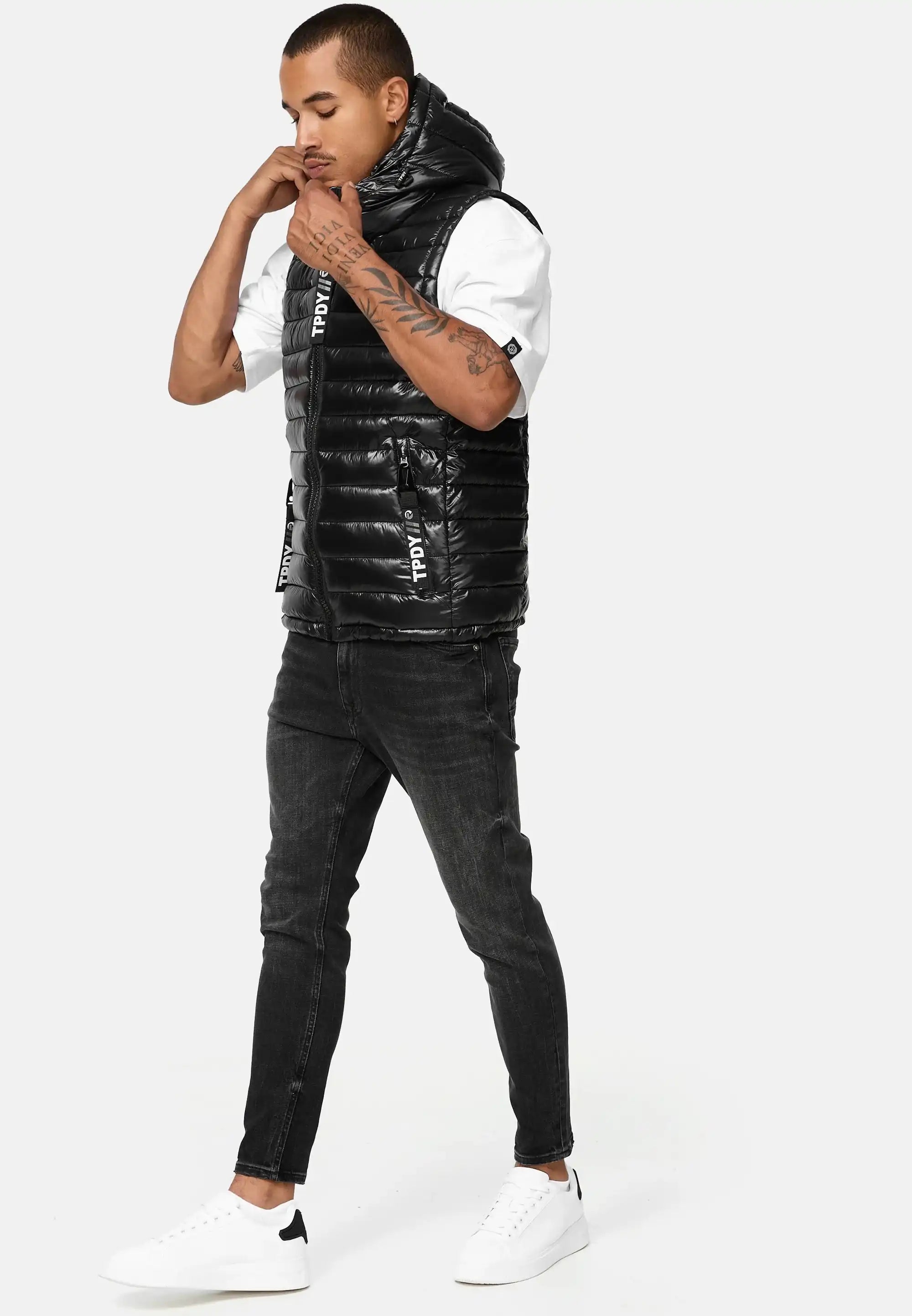 Ein Mann mit Armtattoos steht vor einem schlichten Hintergrund und blickt nach unten, während er die Kapuze seiner Trueprodigy Jared Herrenweste mit Glanz und Kapuze zurechtrückt. Er trägt ein weißes T-Shirt, schwarze Jeans und weiße Turnschuhe.