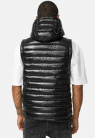 Von hinten ist eine Person zu sehen, die eine glänzende schwarze Trueprodigy Jared Herrenweste mit Glanz und Kapuze über einem weißen kurzärmeligen T-Shirt und dunklen Jeans trägt und auf dem linken Unterarm eine Tätowierung hat.