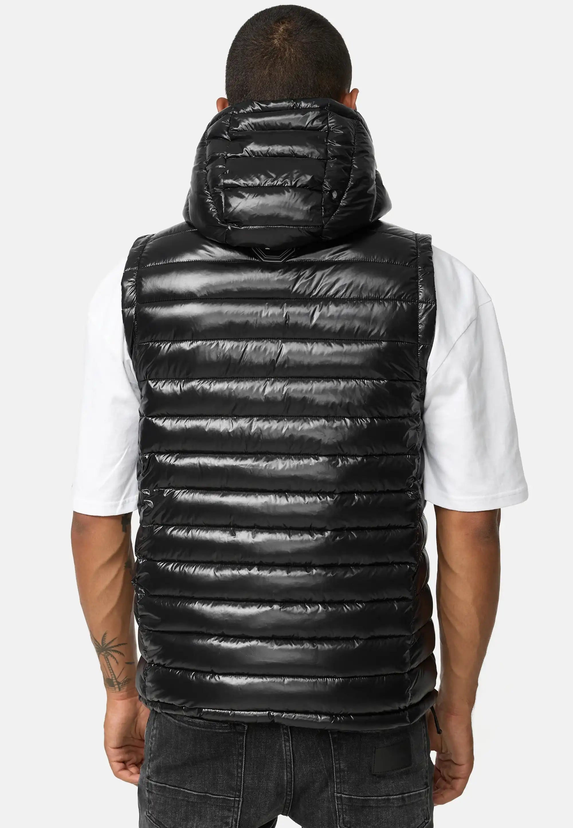 Von hinten ist eine Person zu sehen, die eine glänzende schwarze Trueprodigy Jared Herrenweste mit Glanz und Kapuze über einem weißen kurzärmeligen T-Shirt und dunklen Jeans trägt und auf dem linken Unterarm eine Tätowierung hat.