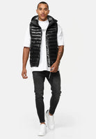 Ein Mann in einer glänzenden schwarzen Trueprodigy Jared Herrenweste mit Glanz und Kapuze über einem weißen T-Shirt, schwarzen Skinny Jeans und weißen Turnschuhen geht vor einem weißen Hintergrund nach vorne. Er hat kurze Haare, eine Tätowierung auf dem linken Arm und einen neutralen Gesichtsausdruck.