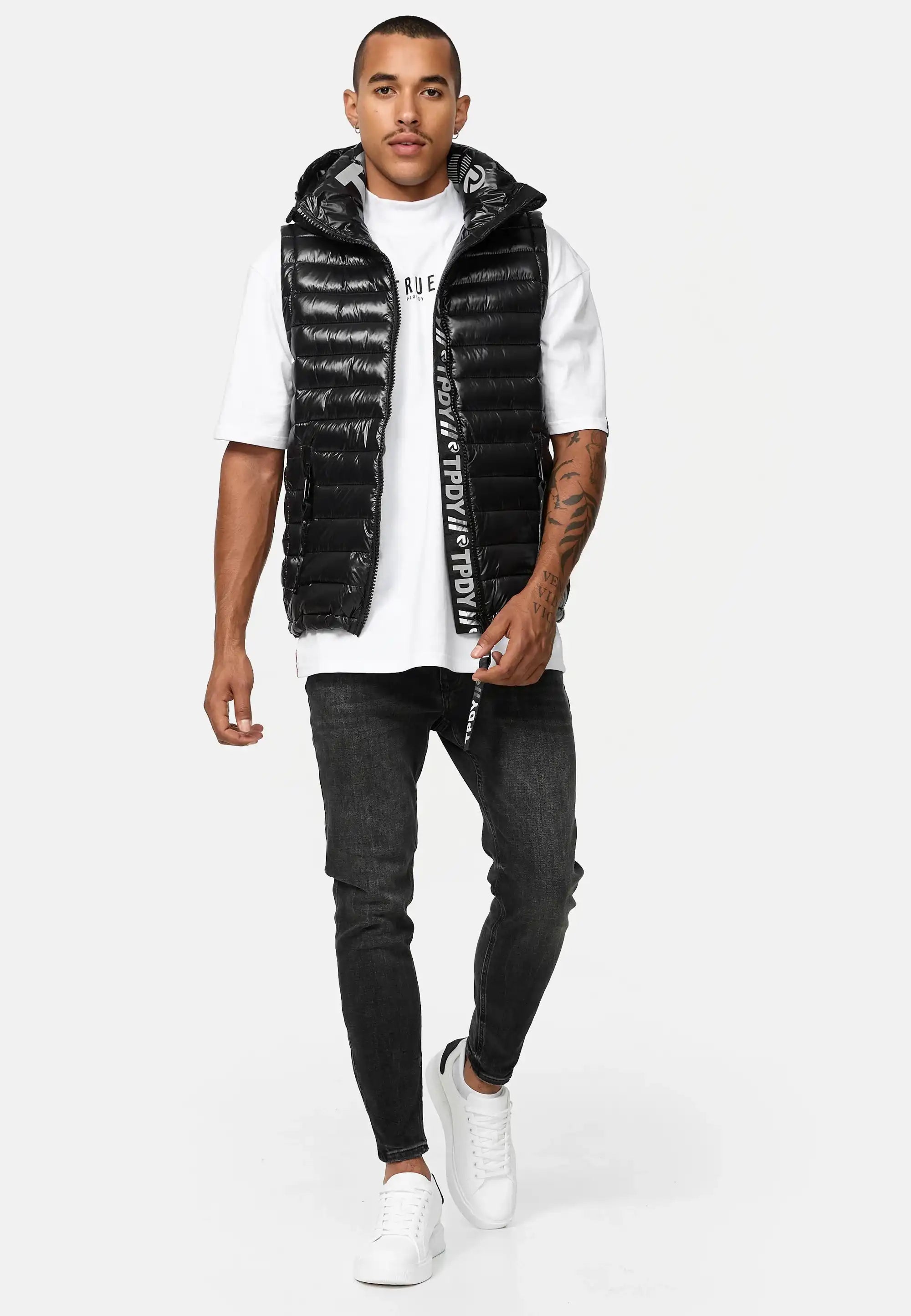 Ein Mann in einer glänzenden schwarzen Trueprodigy Jared Herrenweste mit Glanz und Kapuze über einem weißen T-Shirt, schwarzen Skinny Jeans und weißen Turnschuhen geht vor einem weißen Hintergrund nach vorne. Er hat kurze Haare, eine Tätowierung auf dem linken Arm und einen neutralen Gesichtsausdruck.
