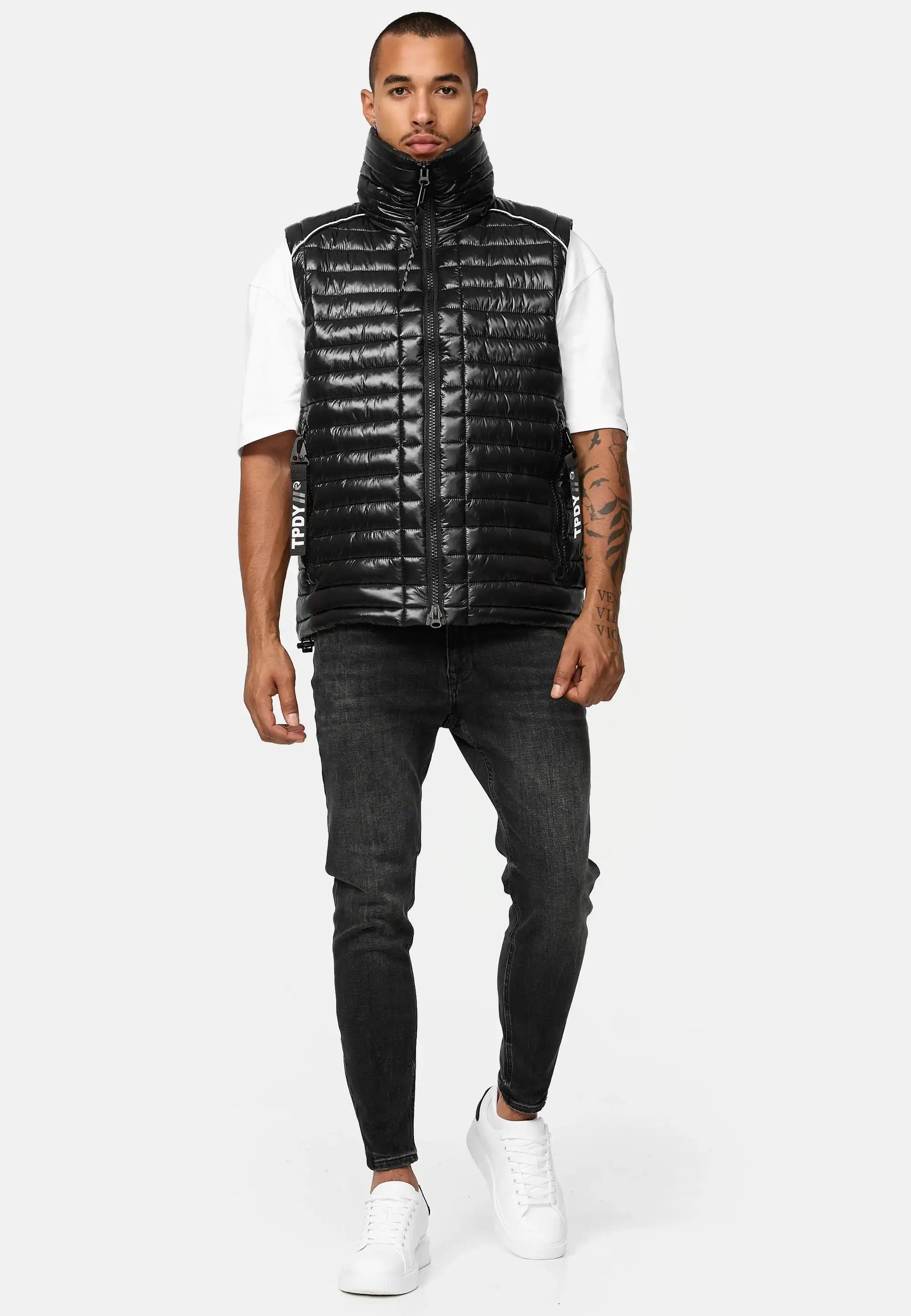 Ein Mann, der eine glänzende schwarze Trueprodigy Dave Herrenweste mit Glanz und Stehkragen über einem weißen kurzärmeligen Hemd, schwarze Skinny Jeans und weiße Turnschuhe trägt, steht vor einem schlichten hellgrauen Hintergrund.