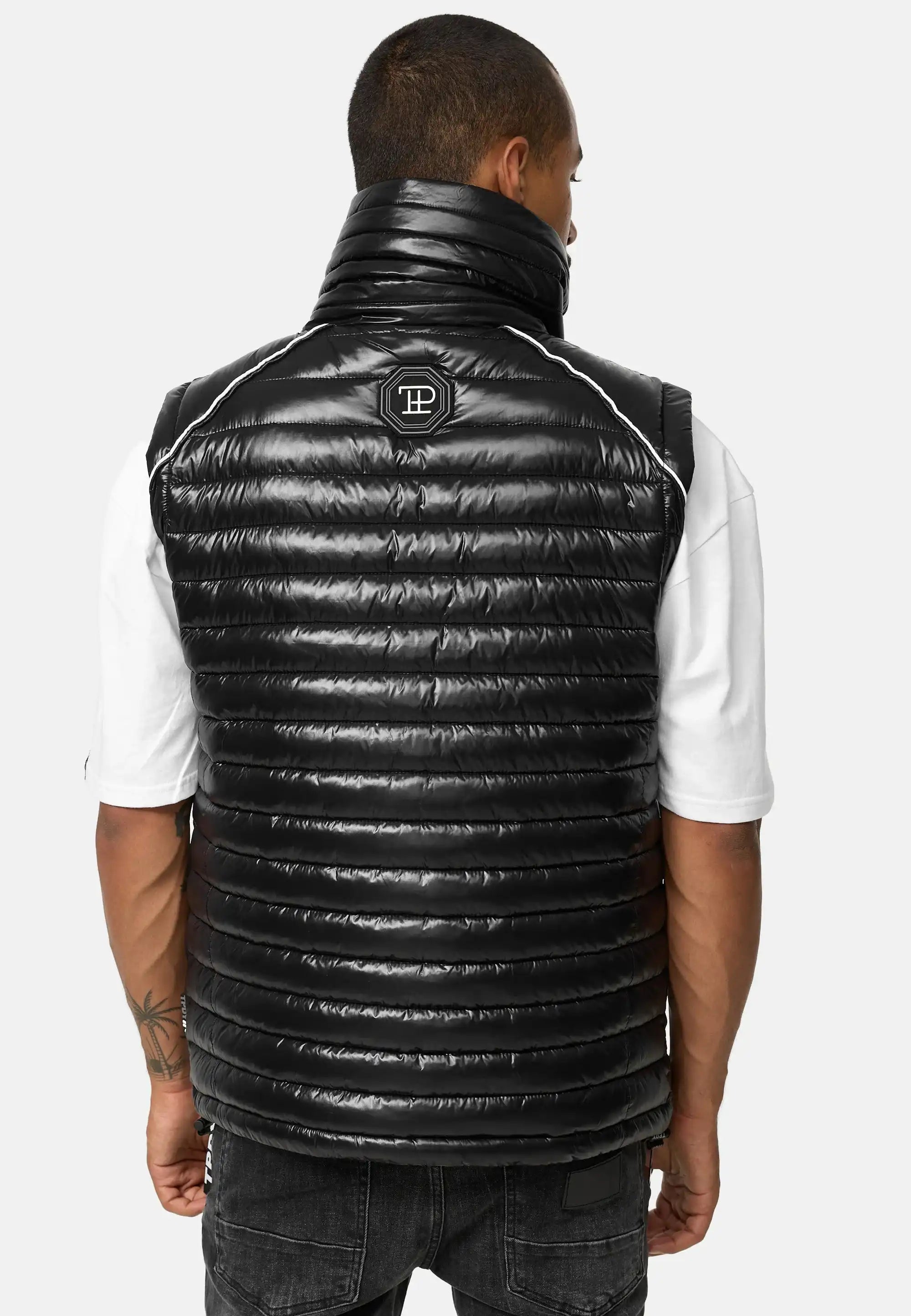 Ein Mann blickt von sich weg und trägt die trueprodigy Dave Herrenweste mit Glanz und Stehkragen mit weißem Logo auf dem oberen Rücken über einem weißen kurzärmeligen Hemd und einer schwarzen Jeans; die Weste ist horizontal gesteppt und hat einen hohen Kragen.