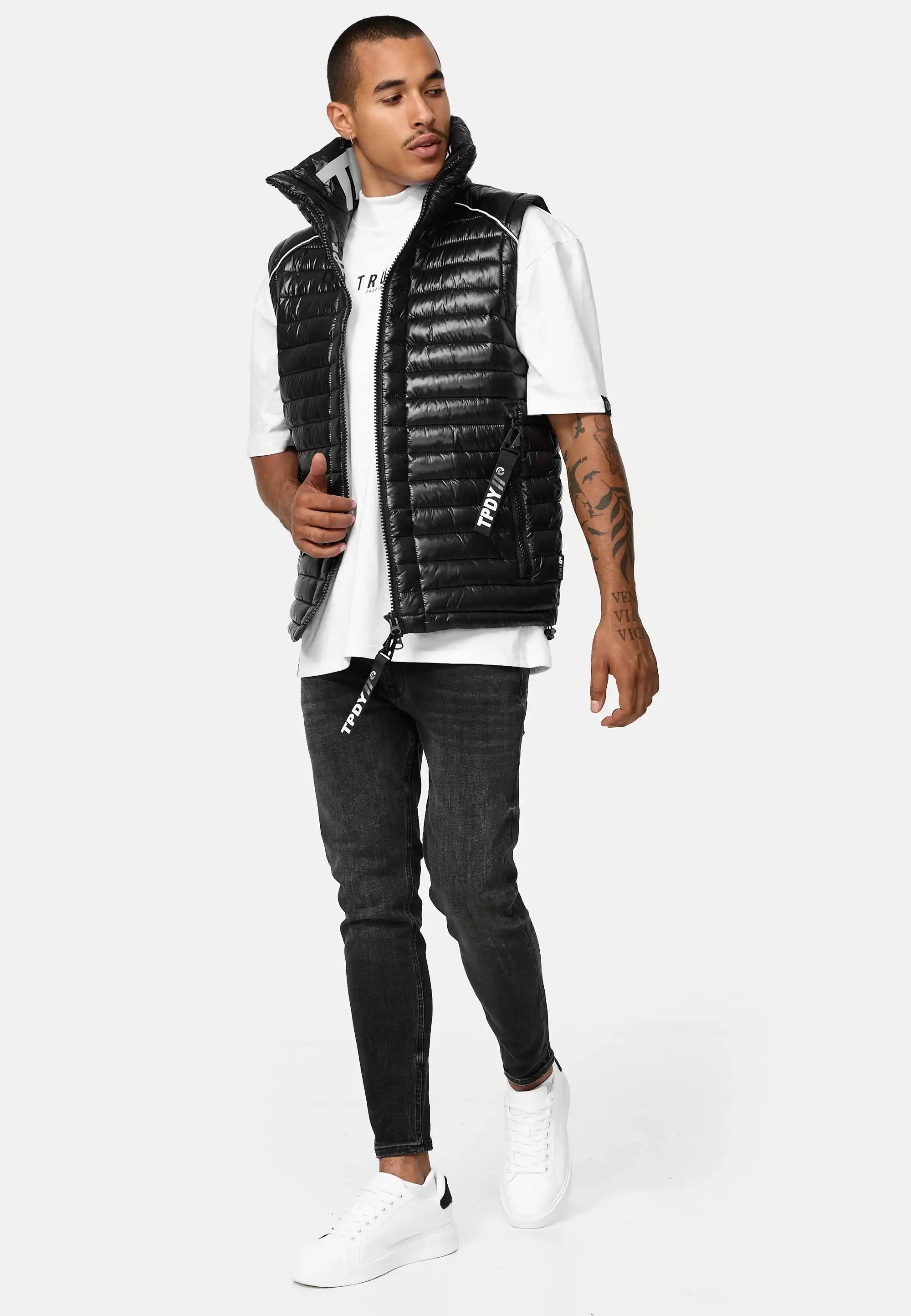 Ein Mann mit kurzen Haaren und sichtbaren Unterarmtattoos steht vor einem schlichten Hintergrund, trägt ein Trueprodigy "Dave Herrenweste mit Glanz und Stehkragen", ein weißes T-Shirt, schwarze Skinny Jeans und weiße Turnschuhe und schaut zur Seite.