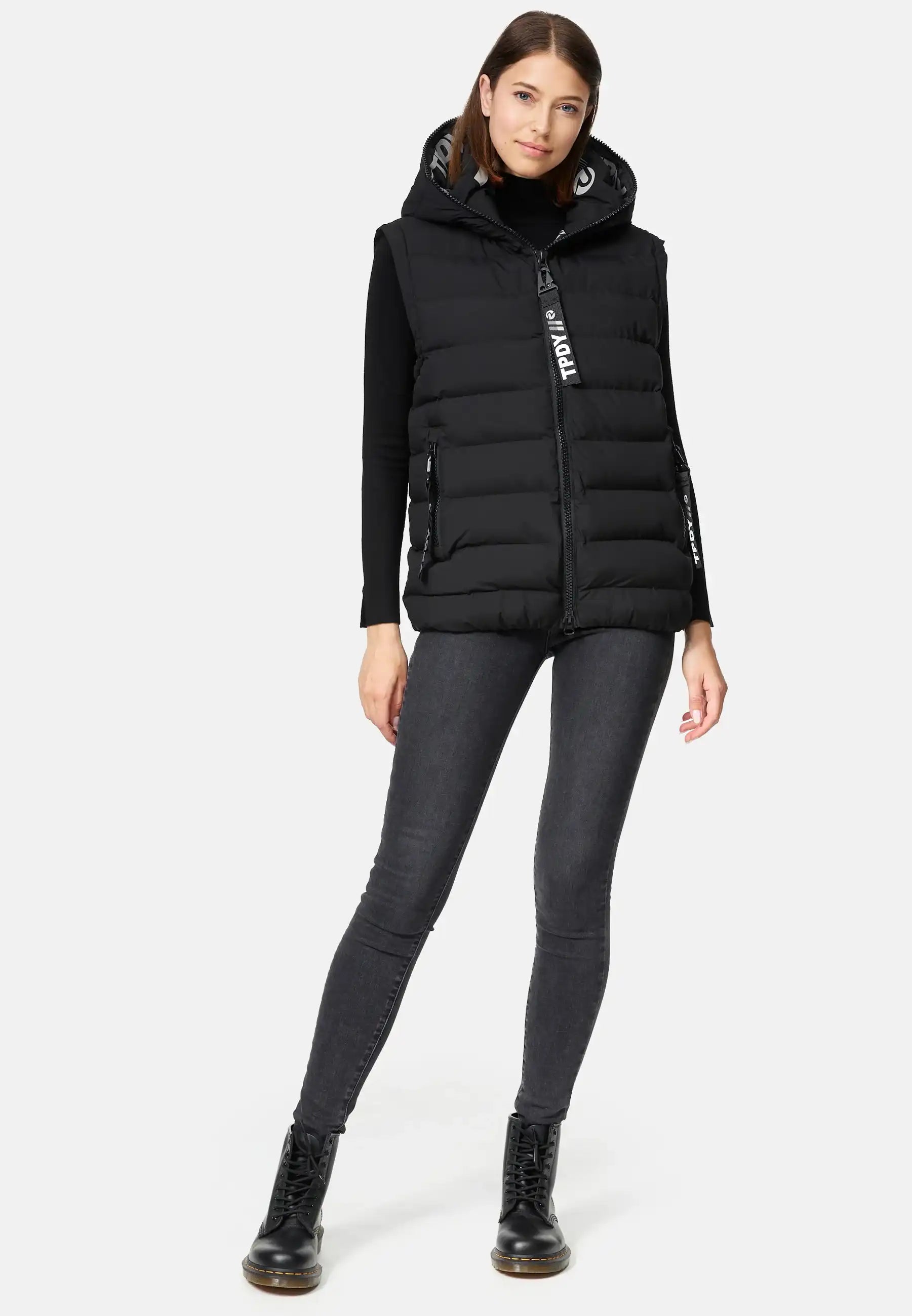 Eine Frau steht vor einem schlichten weißen Hintergrund und trägt eine trueprodigy Matthew F Damenweste mit Kapuze matt, ein schwarzes langärmeliges Oberteil, dunkle Skinny Jeans und schwarze Schnürstiefel.