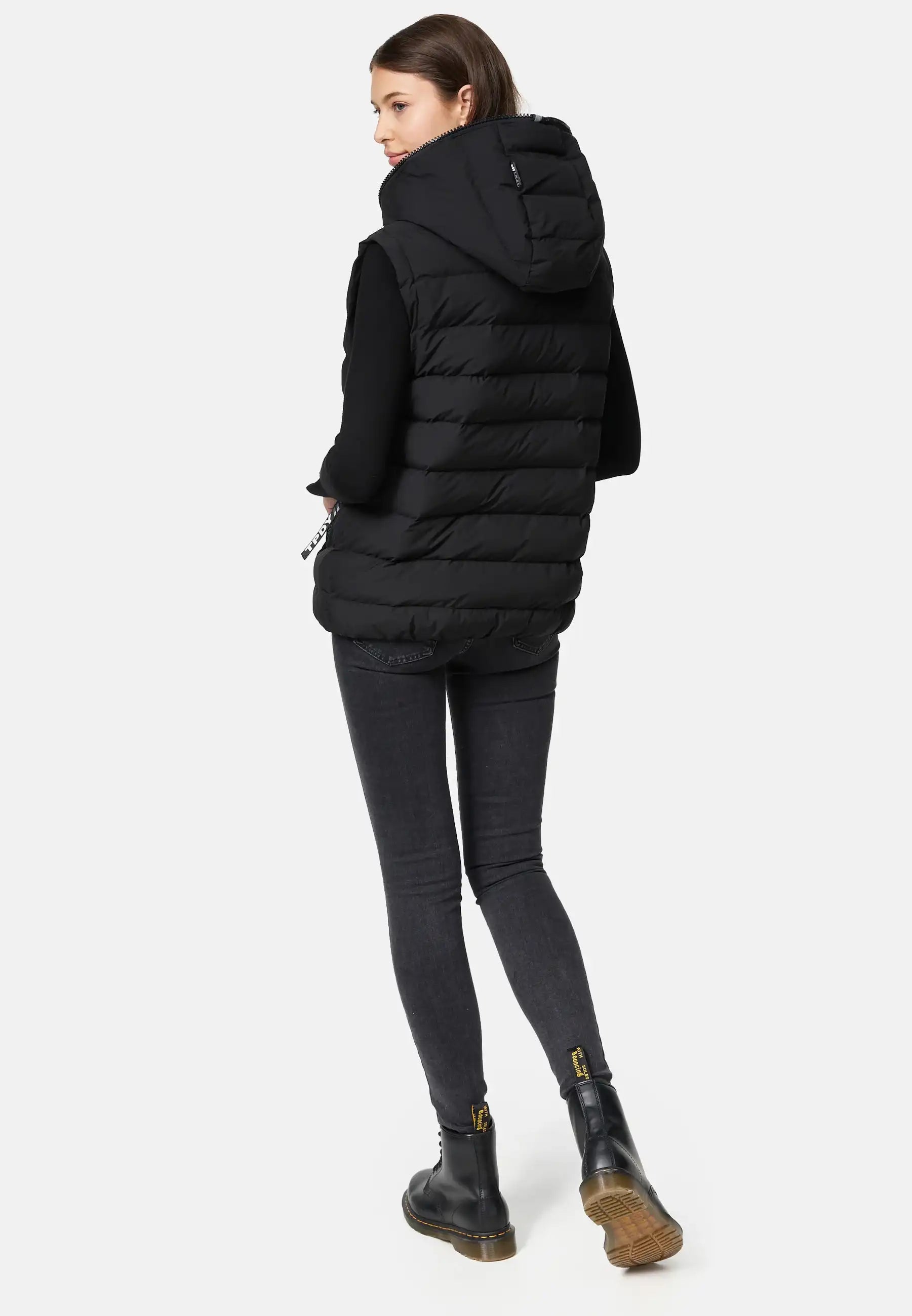 Eine Person steht mit dem Gesicht leicht abgewandt und trägt ein mattes, schwarzes, langärmeliges Oberteil, dunkle Skinny-Jeans und schwarze Ankle Boots mit gelben Laschen vor einem hellgrauen Hintergrund.