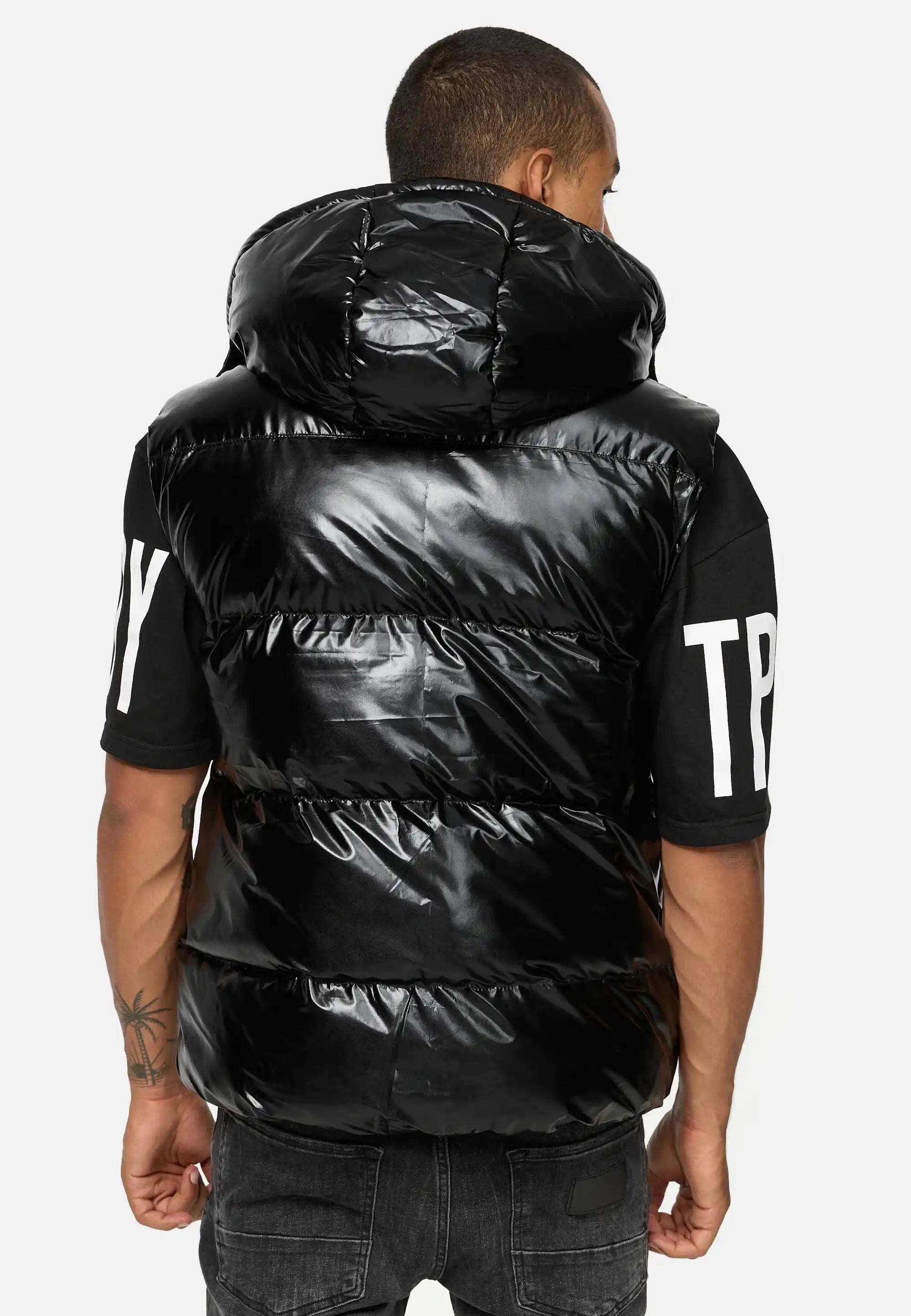 Von hinten ist eine Person zu sehen, die eine schwarze Darko Steppweste glänzend mit Kapuze von trueprodigy über einem schwarzen T-Shirt mit weißem Schriftzug an den Ärmeln trägt, gepaart mit einer dunklen Jeans.