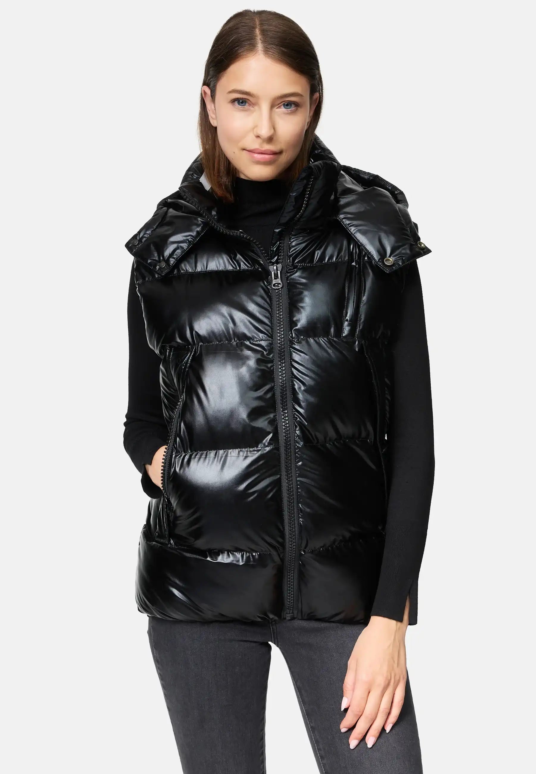 Eine Frau mit glattem braunem Haar trägt die trueprodigy Darko F Weste glänzend mit Kapuze über einem schwarzen Rollkragenpullover und grauen Jeans, eine Hand in der Westentasche, vor einem schlichten hellen Hintergrund.