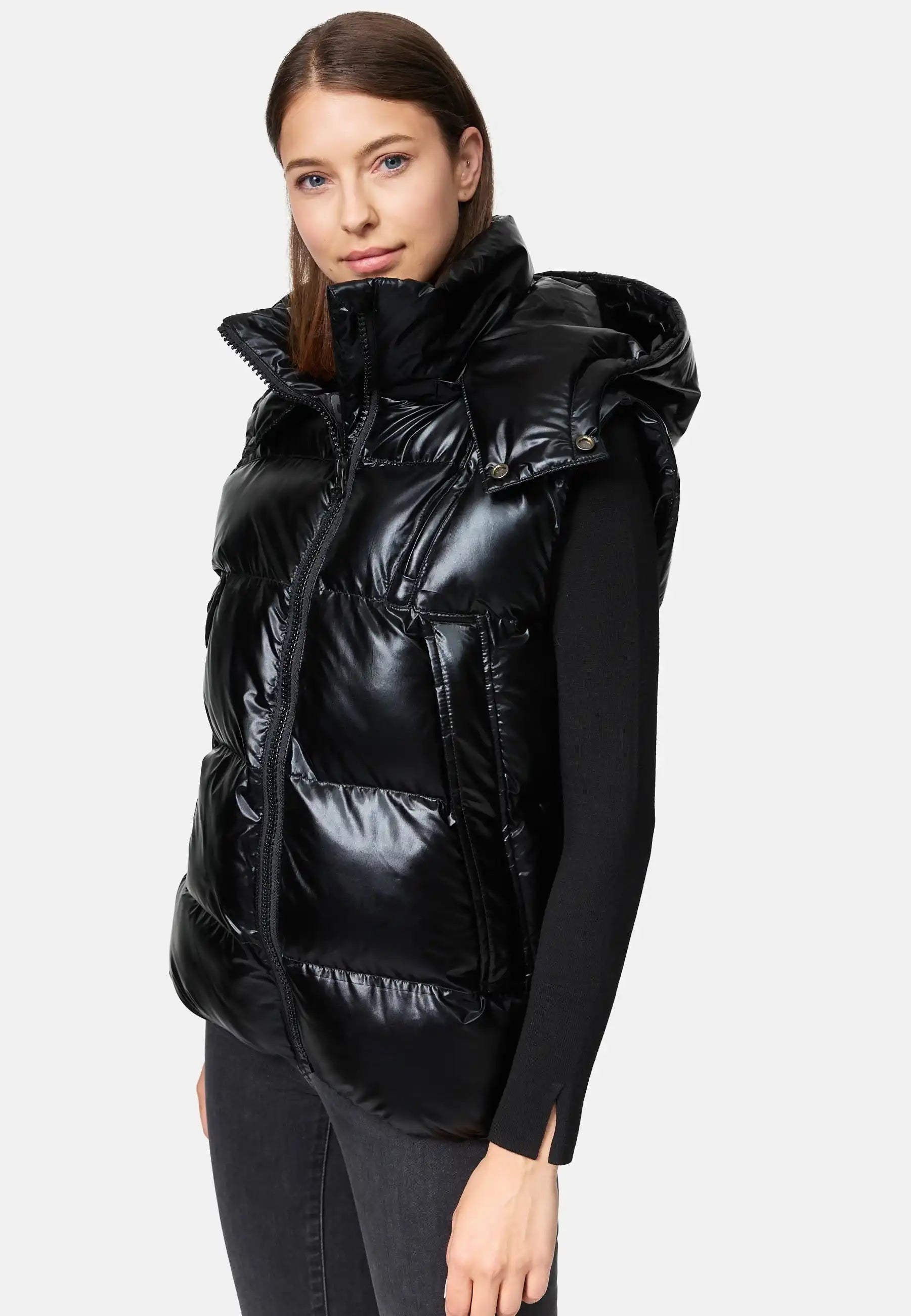 Eine Person, die die Trueprodigy Darko F Weste glänzend mit Kapuze über einem schwarzen langärmeligen Oberteil und dunklen Jeans trägt, steht vor einem schlichten hellen Hintergrund.