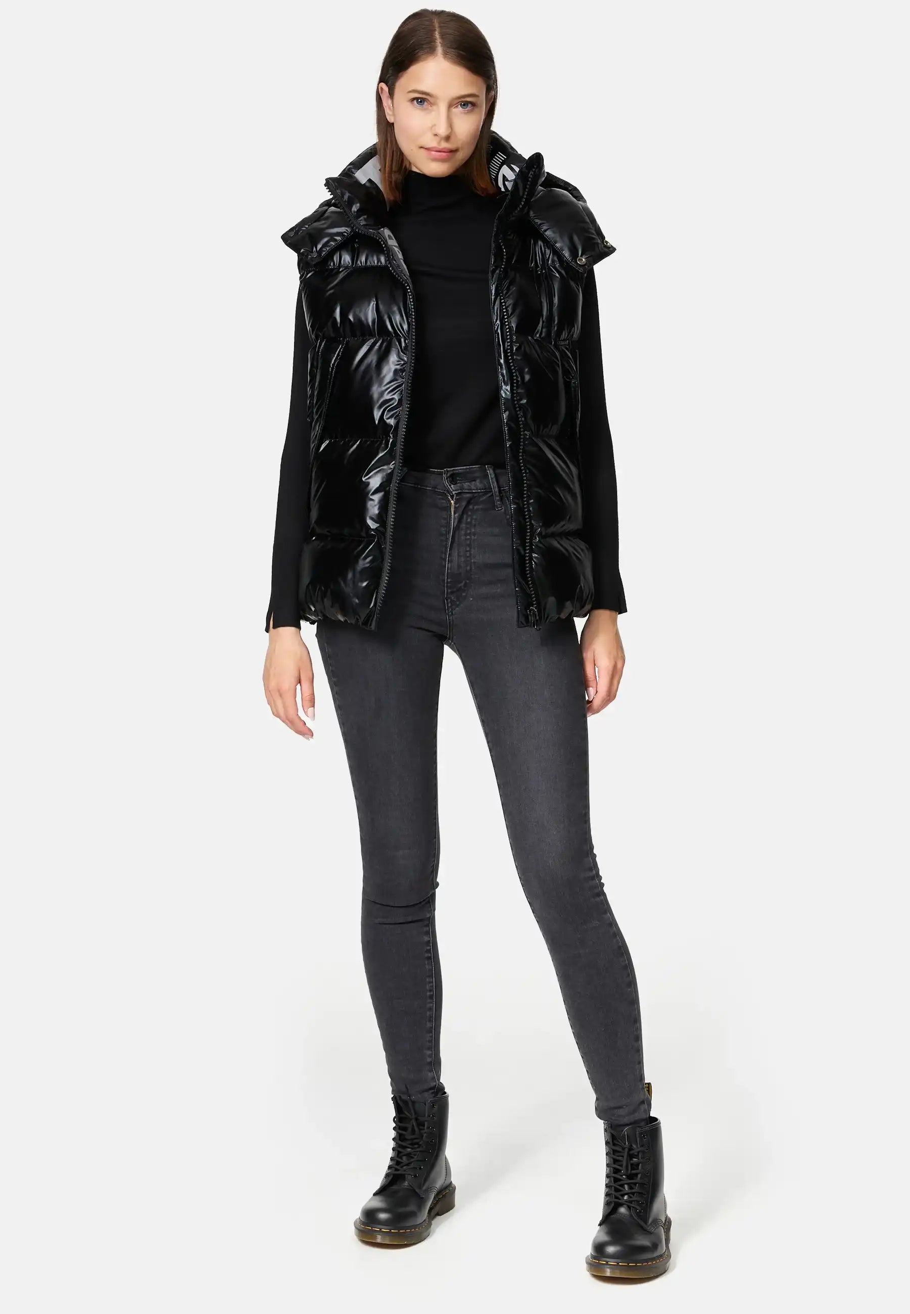 Eine Frau steht mit dem Gesicht nach vorne und trägt die Trueprodigy Darko F Weste glänzend mit Kapuze in Schwarz, einen schwarzen Rollkragenpullover, dunkelgraue Skinny Jeans und schwarze Schnürstiefel vor einem schlichten hellen Hintergrund.